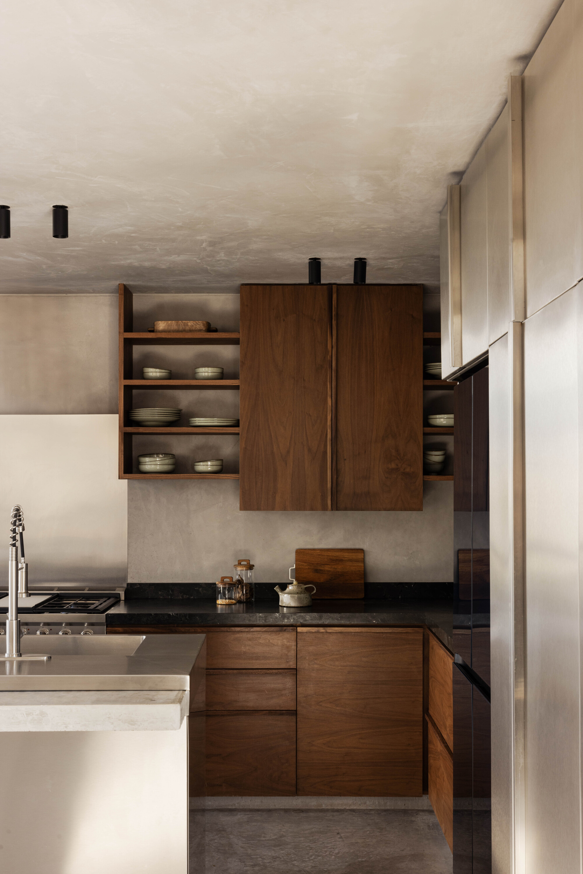 Kitchen corner with walnut cabinets, open shelving of stacked ceramics and a dark stone counter beside stainless steel panels in soft daylight