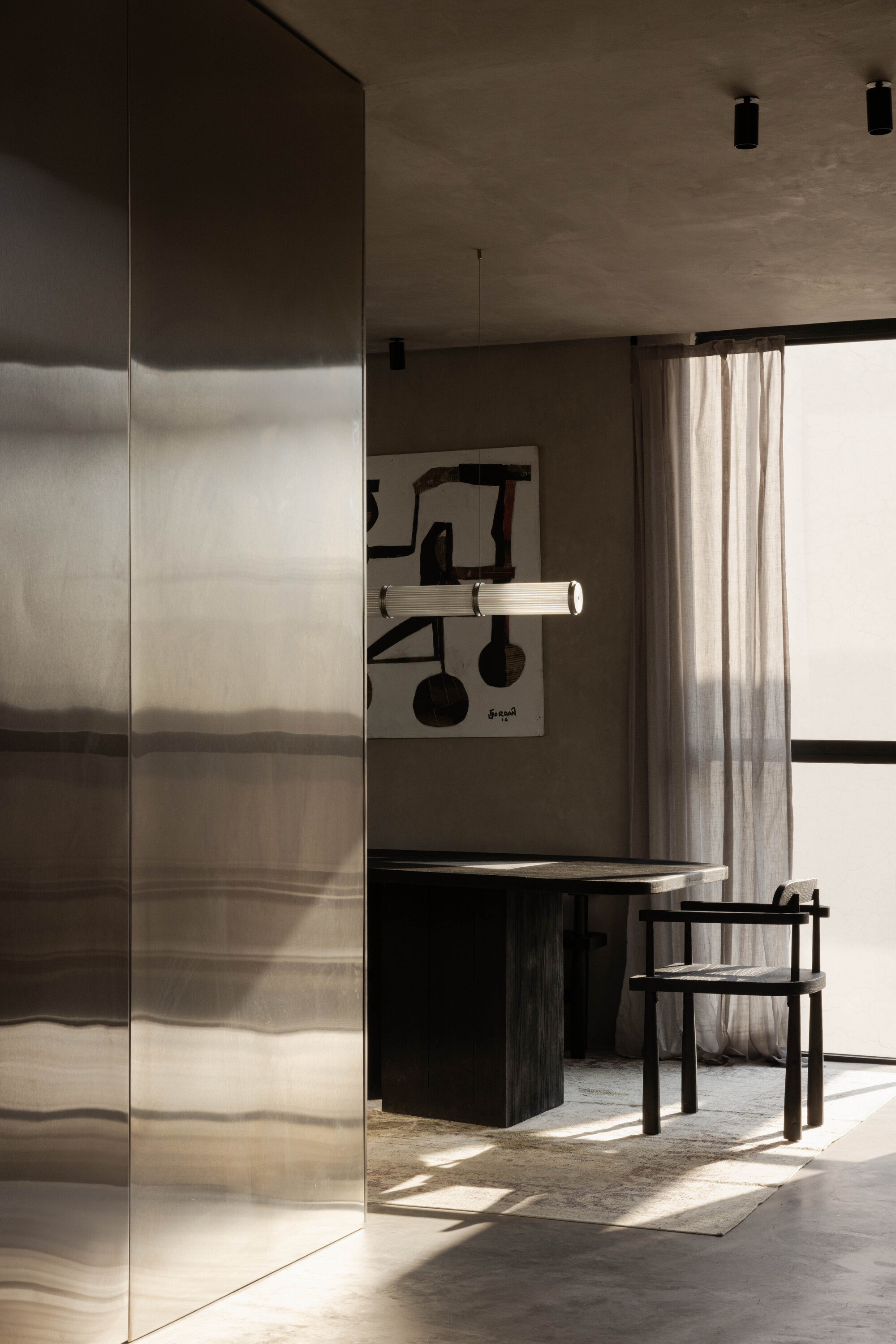 Dining table and black wooden chair seen past a polished stainless steel wall, with sheer curtains and sunlight across the floor