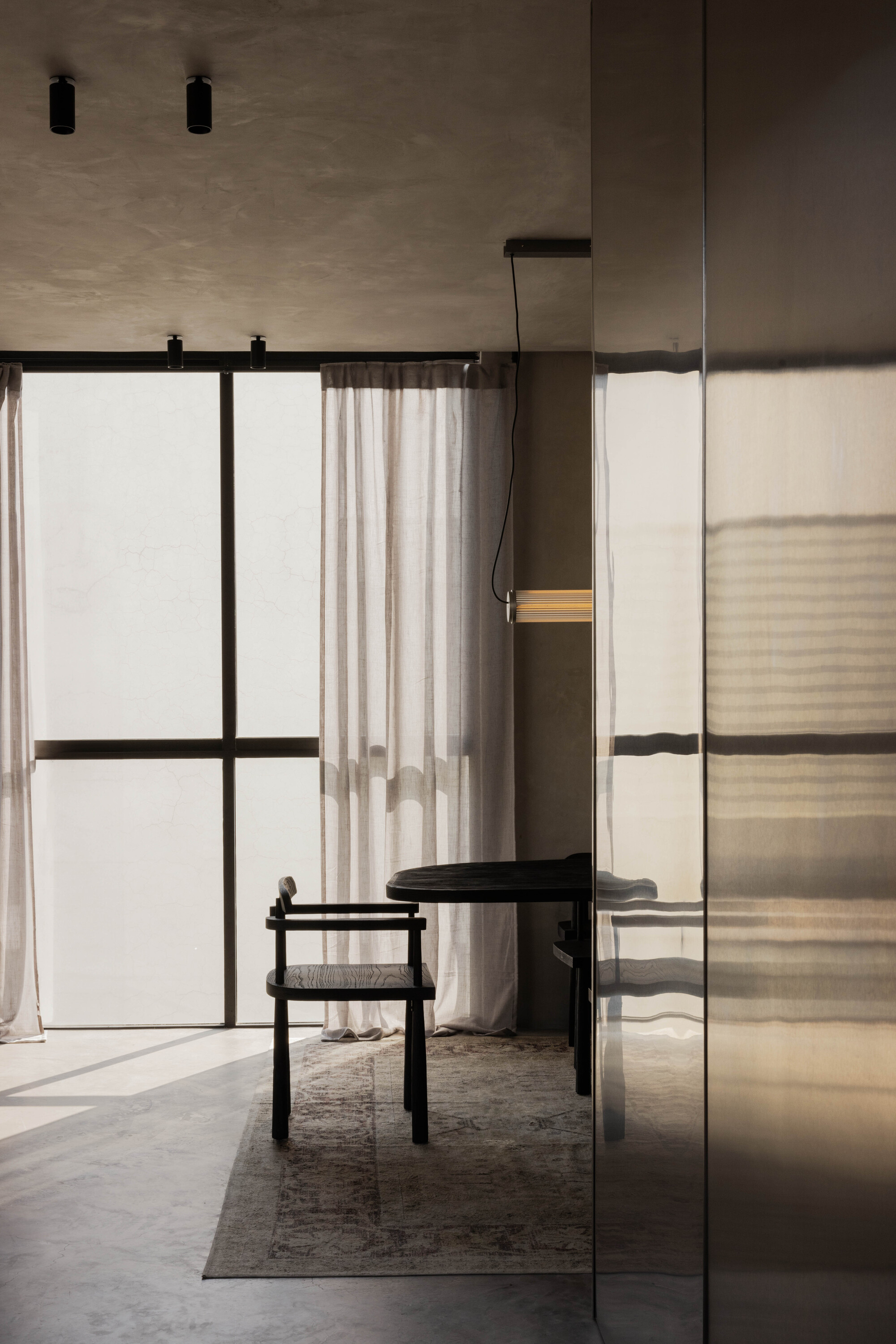 Black wooden chair and round dining table beside floor-to-ceiling glazing with linen curtains, reflected in a steel panel at late afternoon