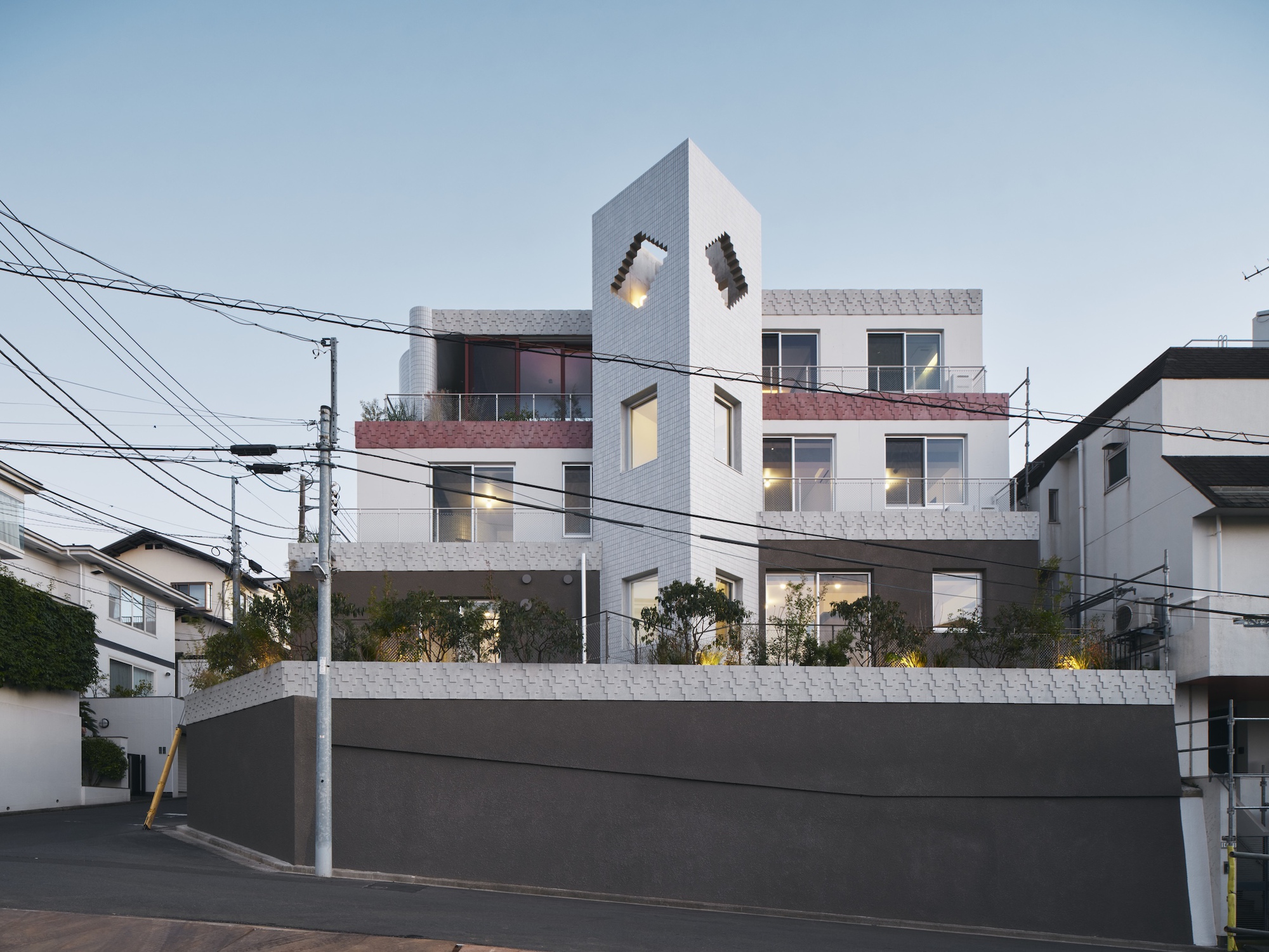 Street-level view of the stepped white tiled block with a tall tower and planted terraces above a dark retaining wall at dusk