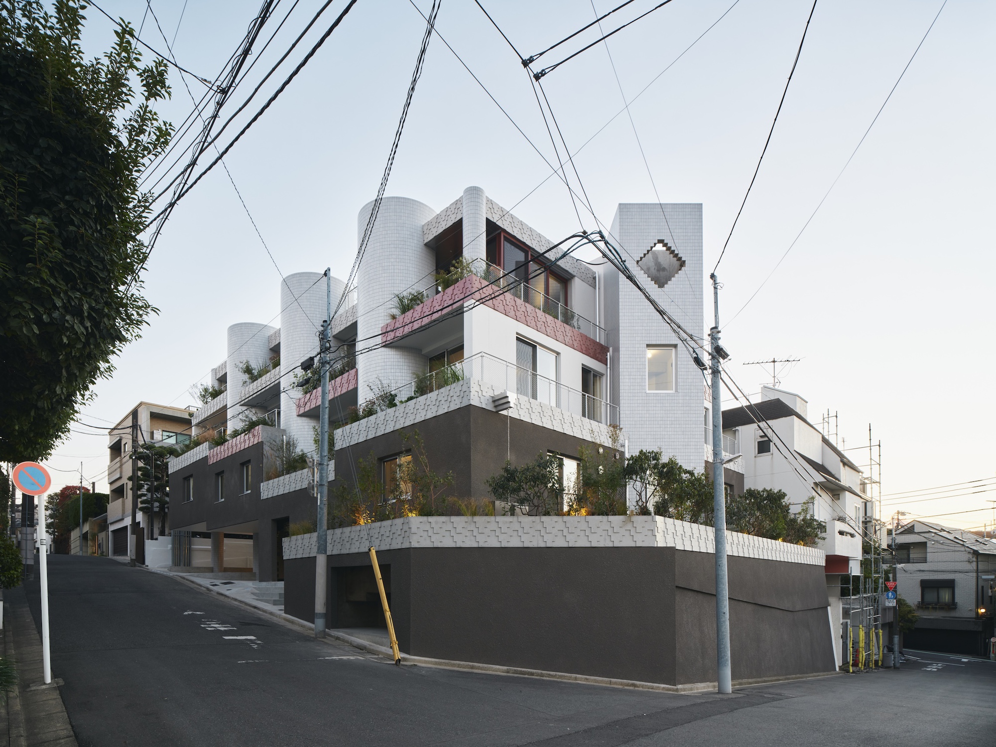 Corner view of terraced volumes with cylindrical tiled towers and planted balconies above a dark base, seen at dusk