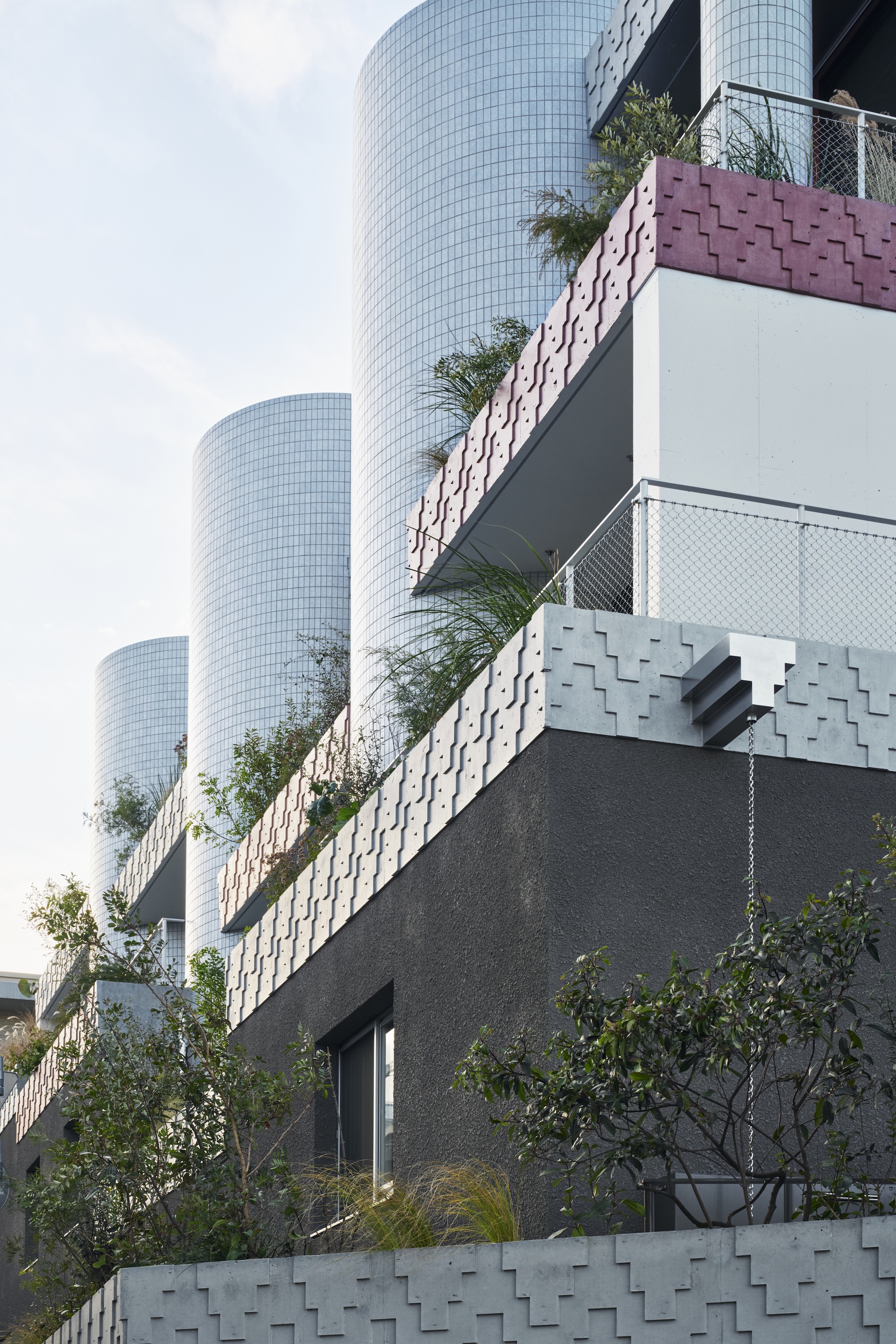 Upward view of cylindrical tiled towers rising behind patterned concrete balcony edges and grasses under a pale sky