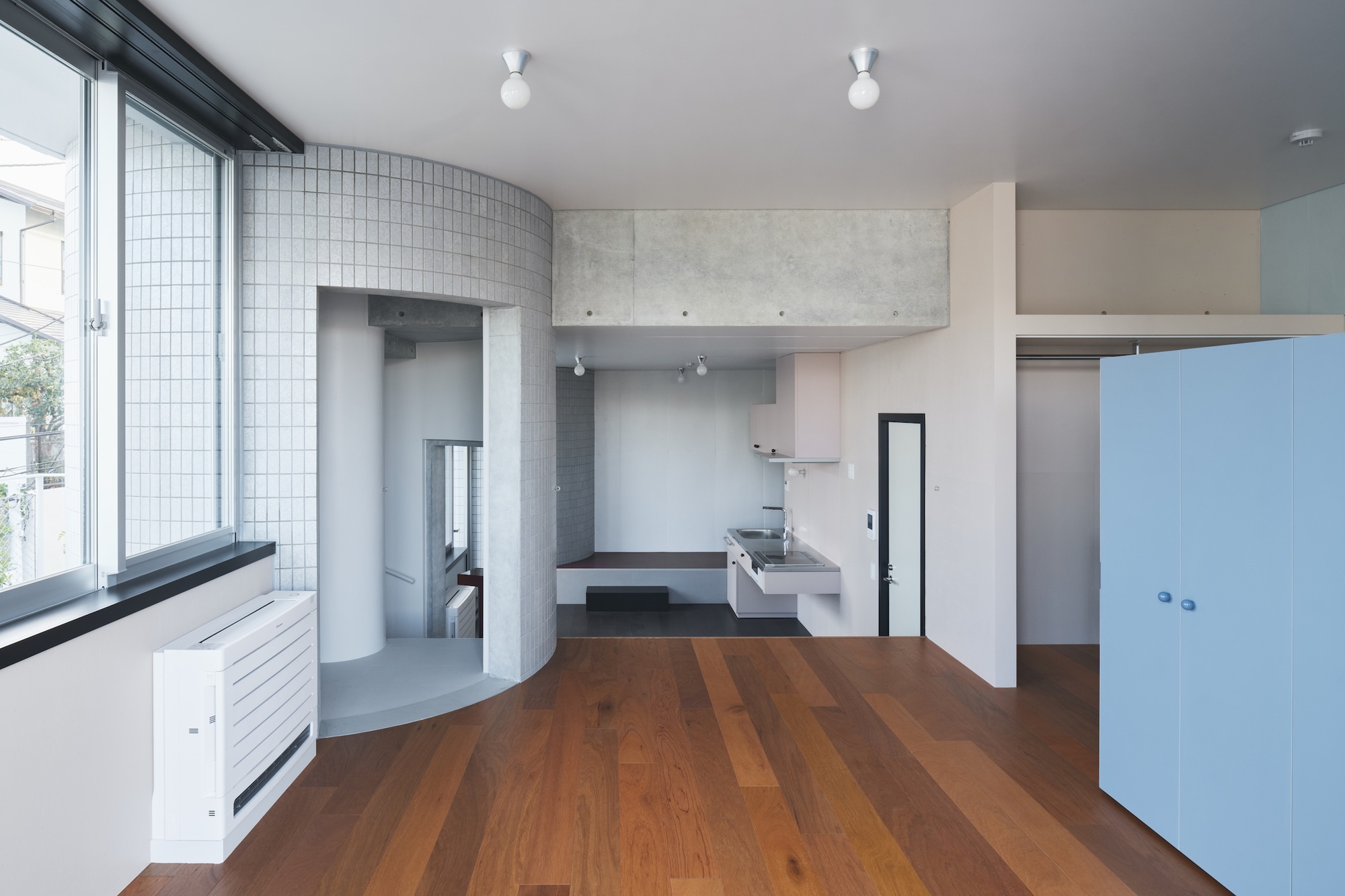 Living space with a curved tiled wall enclosing a stair, a kitchen at the back and a pale blue cabinet on wood flooring