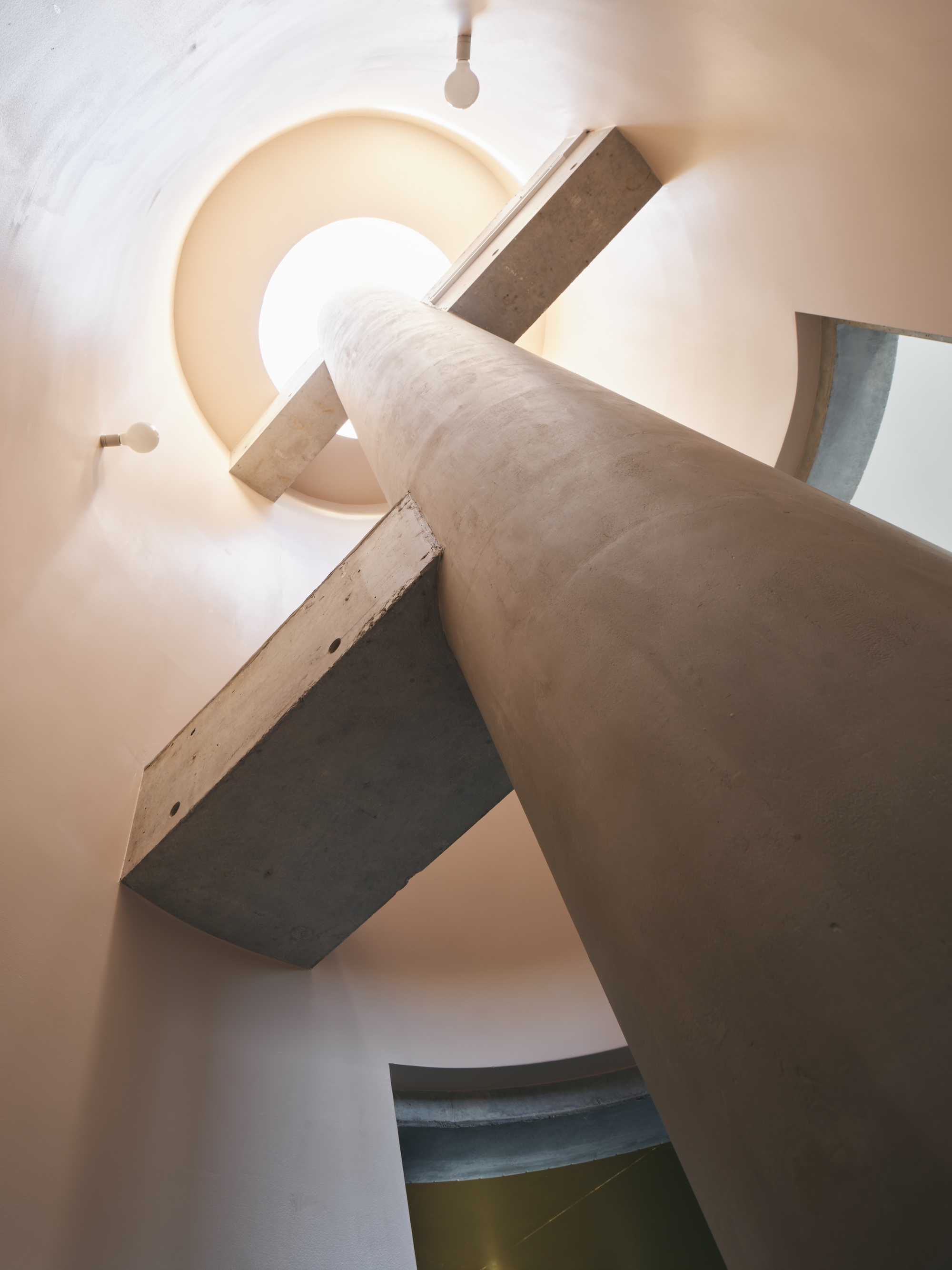Looking up a round stairwell where a concrete column and cantilevered concrete treads rise toward a glowing skylight