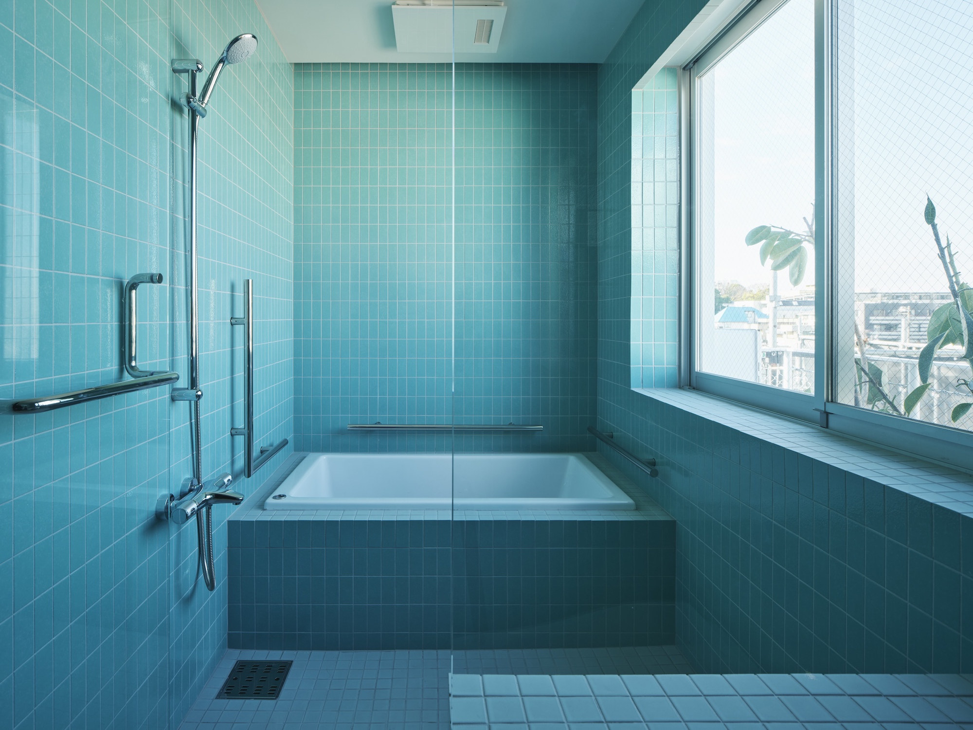Bathroom clad in blue-green square tiles with a sunken tub, handheld shower and a large window onto leafy treetops