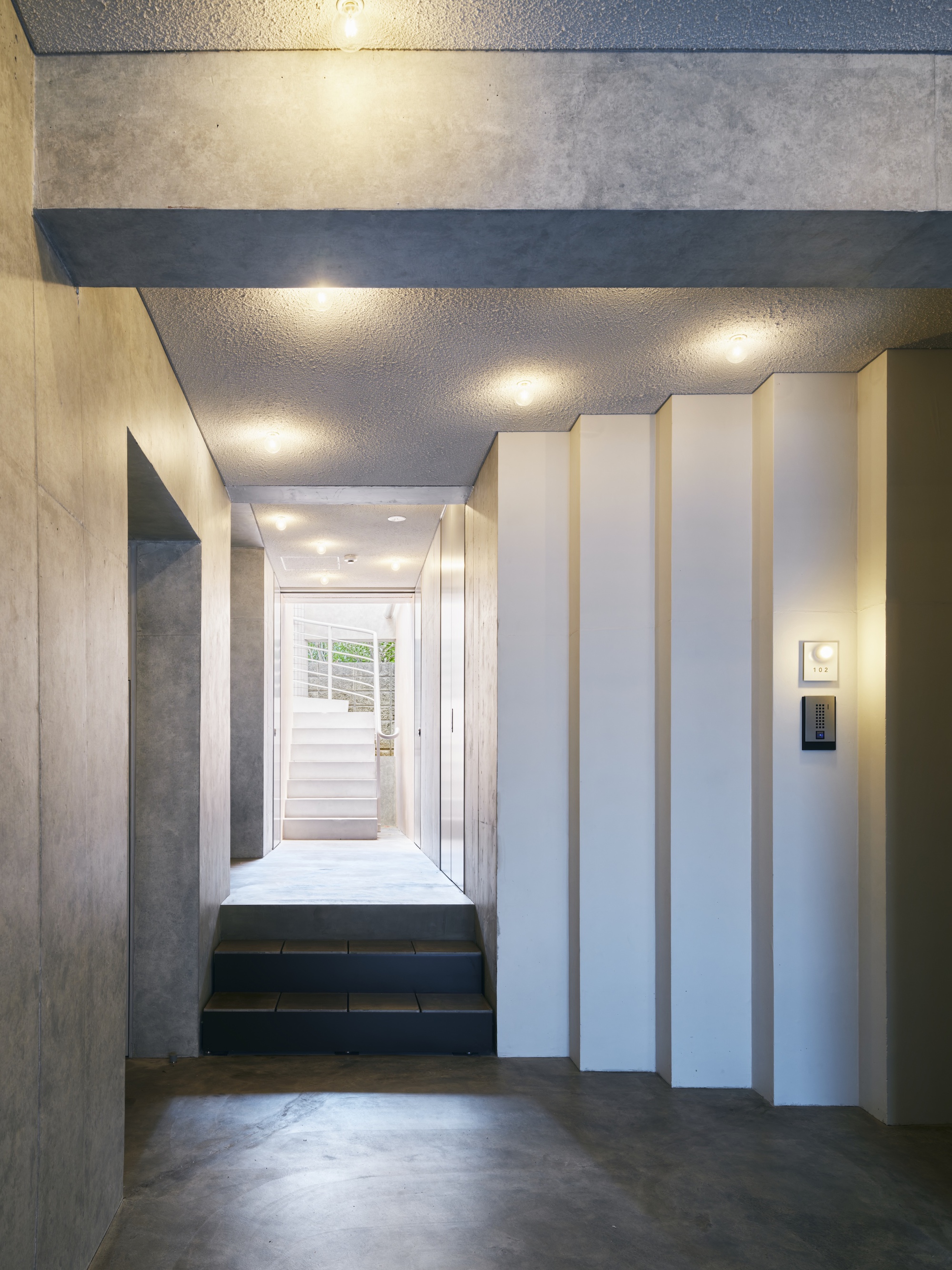 Concrete entry corridor with pleated white wall panels, dark stone steps and ceiling downlights leading toward a bright stair