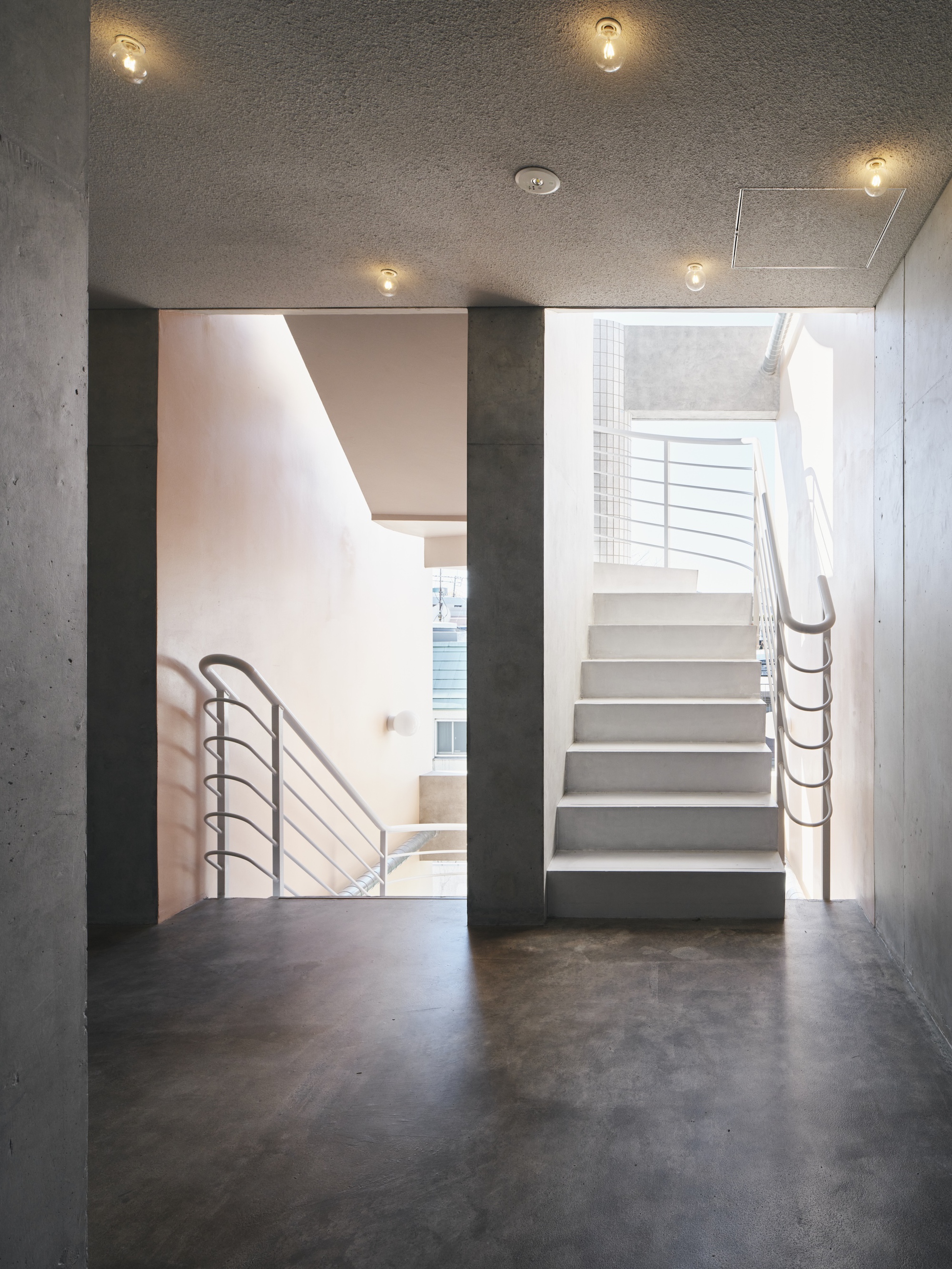 Stairwell landing with white steel railings, a raw concrete column and bare ceiling bulbs, lit by daylight from a side opening