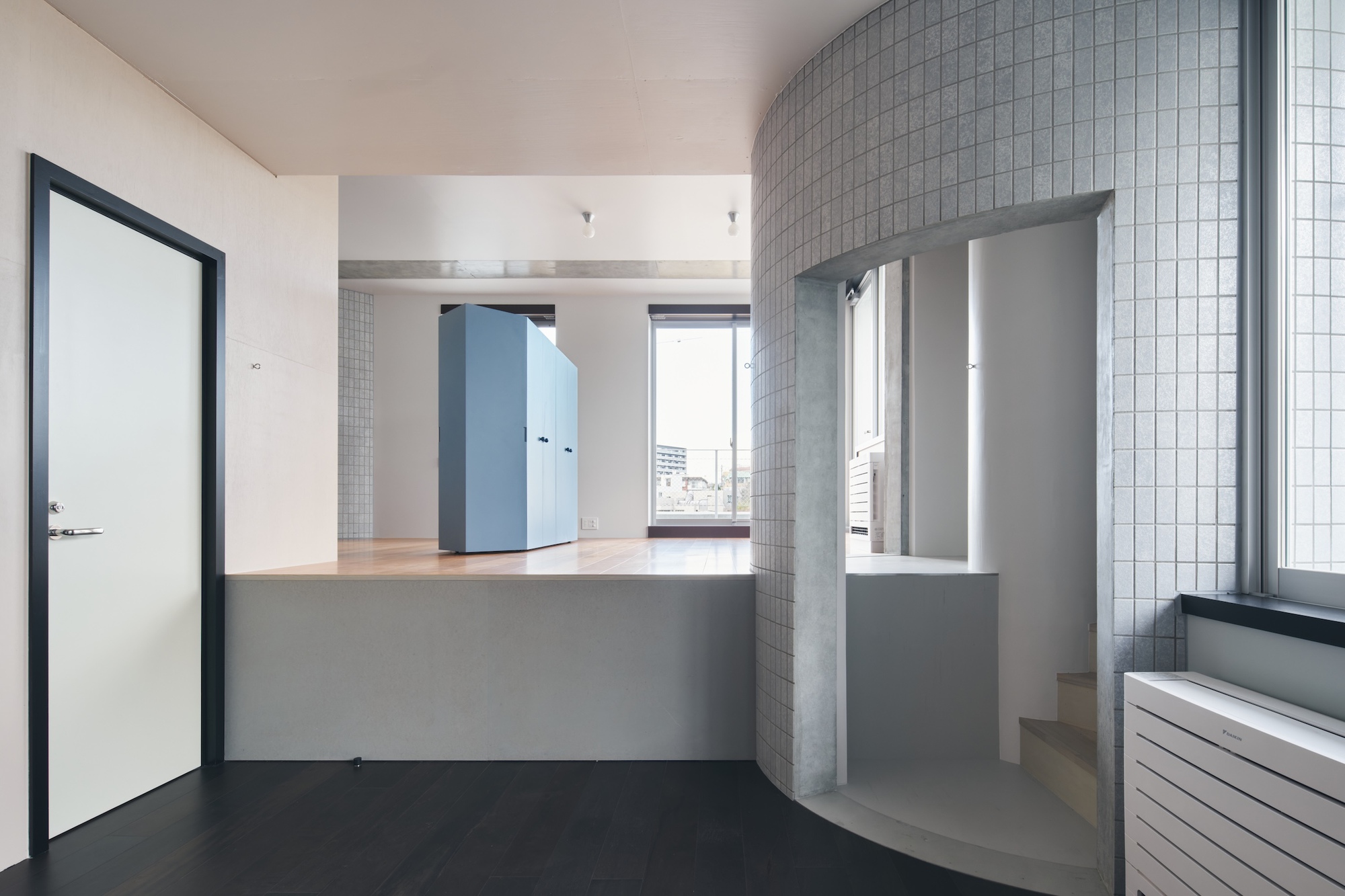 Raised living platform with a pale blue storage cabinet, curved grey tiled wall with arched opening to a stair, and daylight from the window