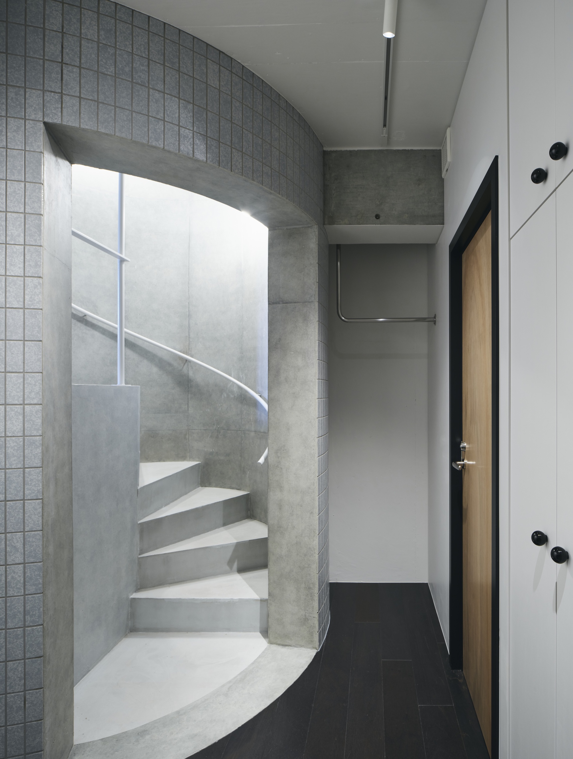 Spiral concrete stair set in a curved tiled alcove beside a timber door and white cabinets, lit from above