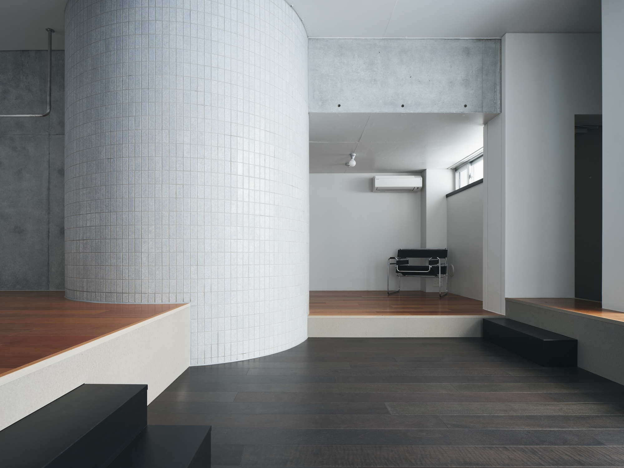 Curved white-tiled wall above a stepped platform, with a black chair against a far wall in soft daylight