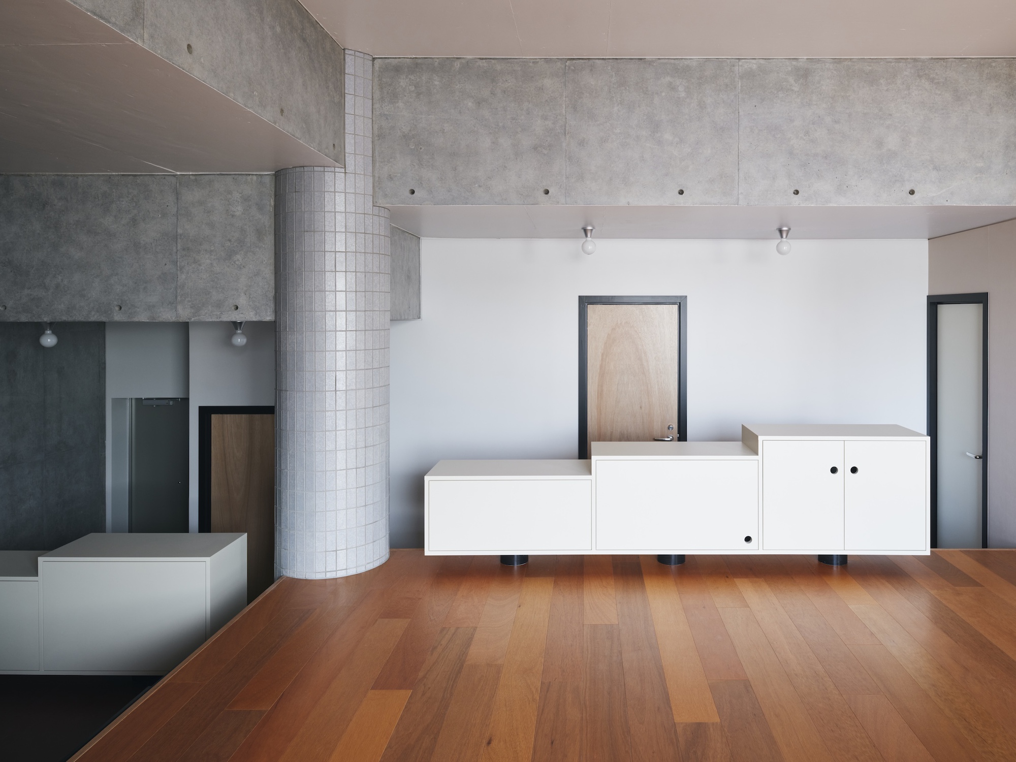 Low white cabinet unit standing on black feet before a timber door, beneath a board-marked concrete ceiling