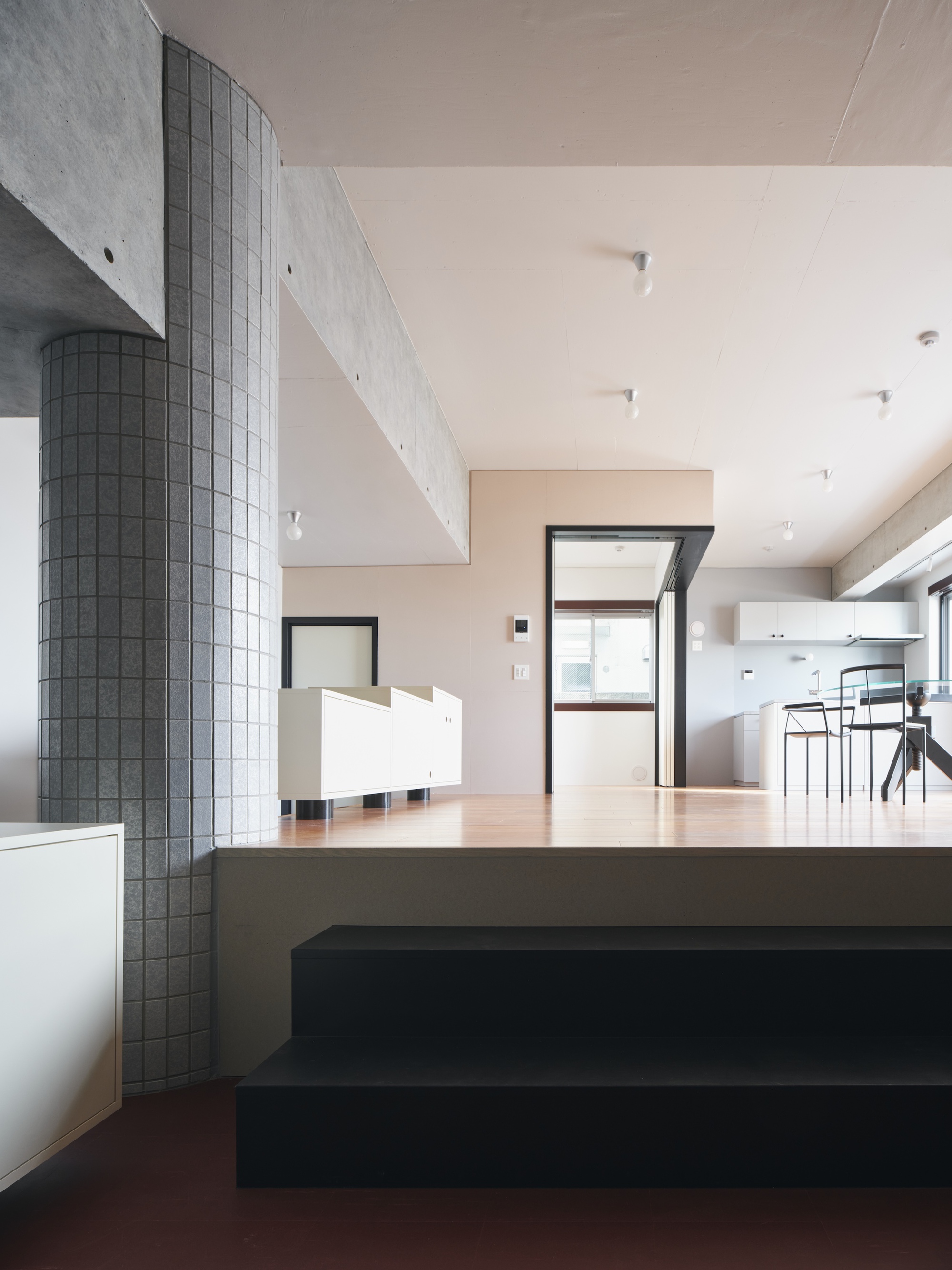 Black stair steps rising to a timber platform beside a grey-tiled column, with the kitchen glowing in daylight beyond