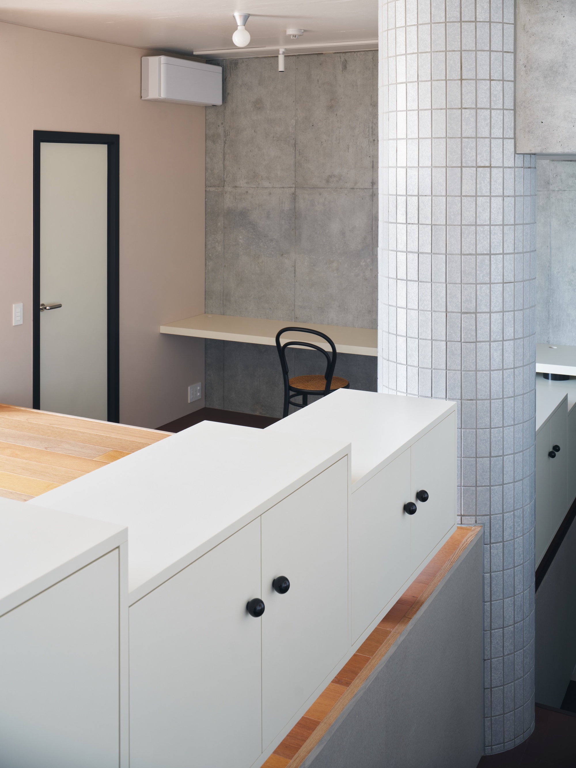 Stepped white cabinets with black knobs beside a tiled column, and a bentwood chair at a concrete wall desk