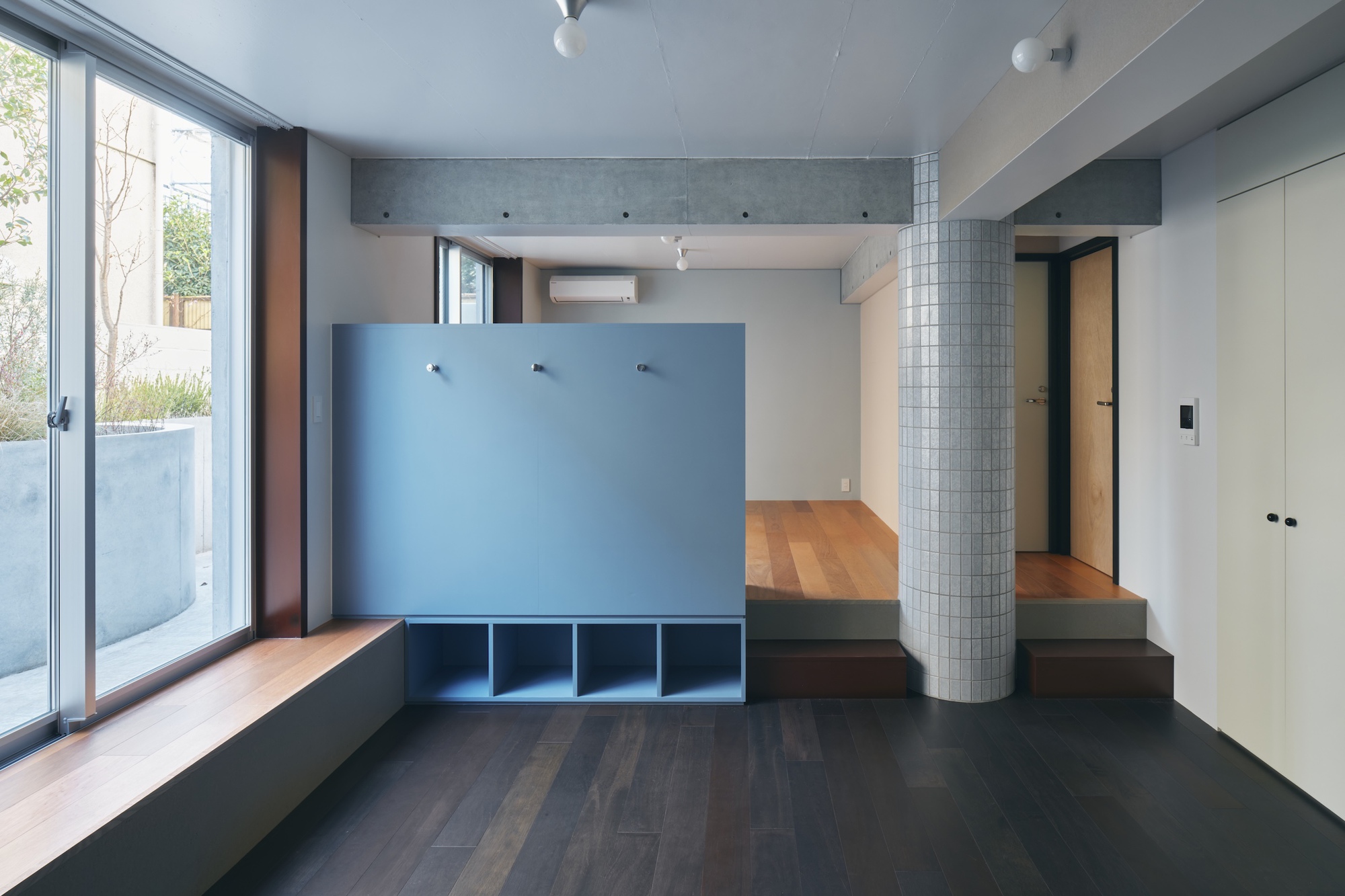 Blue storage partition with cubby shelves and coat hooks beside a tiled column, with a timber window bench in daylight