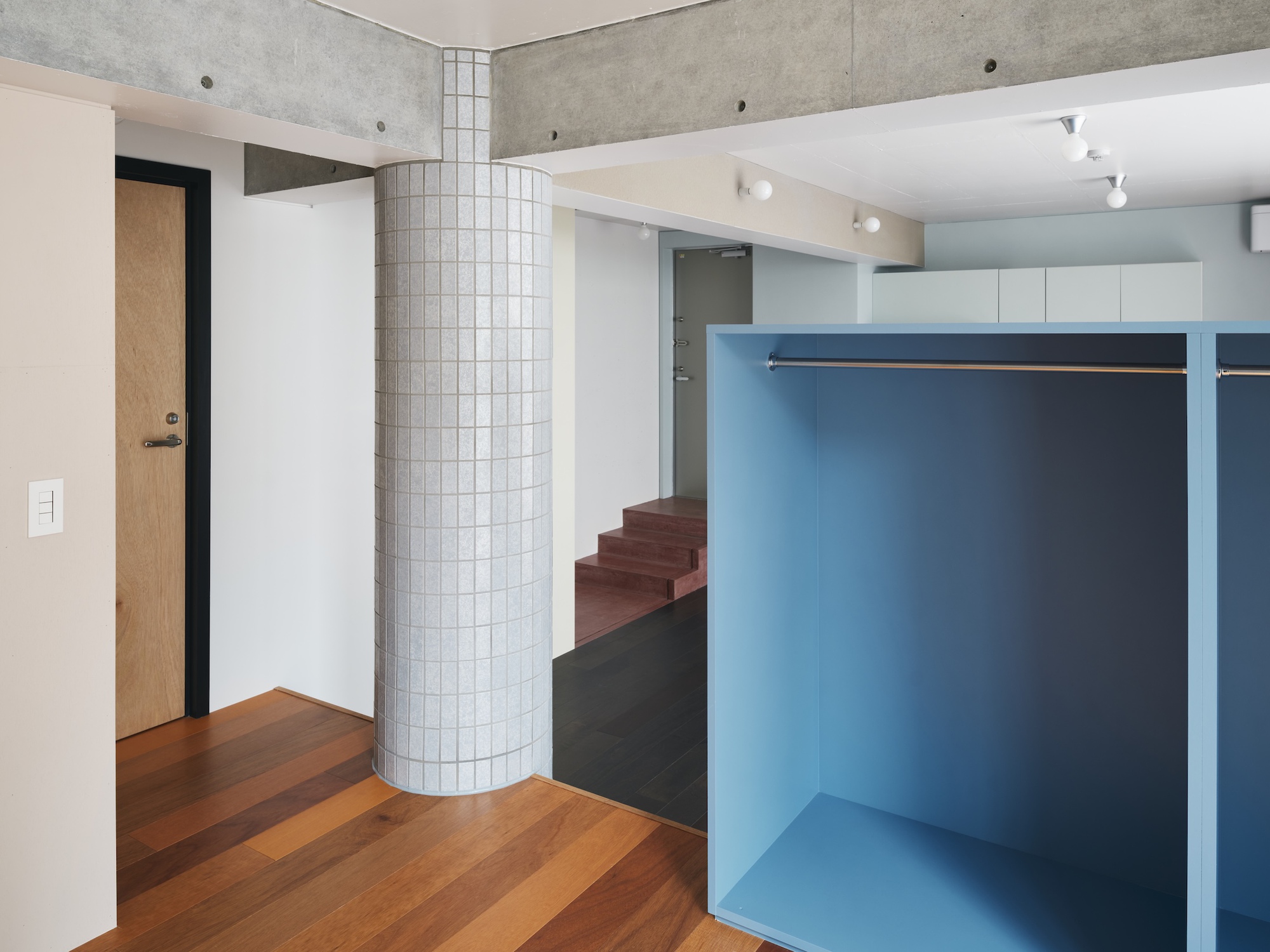 Open blue wardrobe unit beside a grid-tiled column, with exposed concrete beams and red stair treads in the background