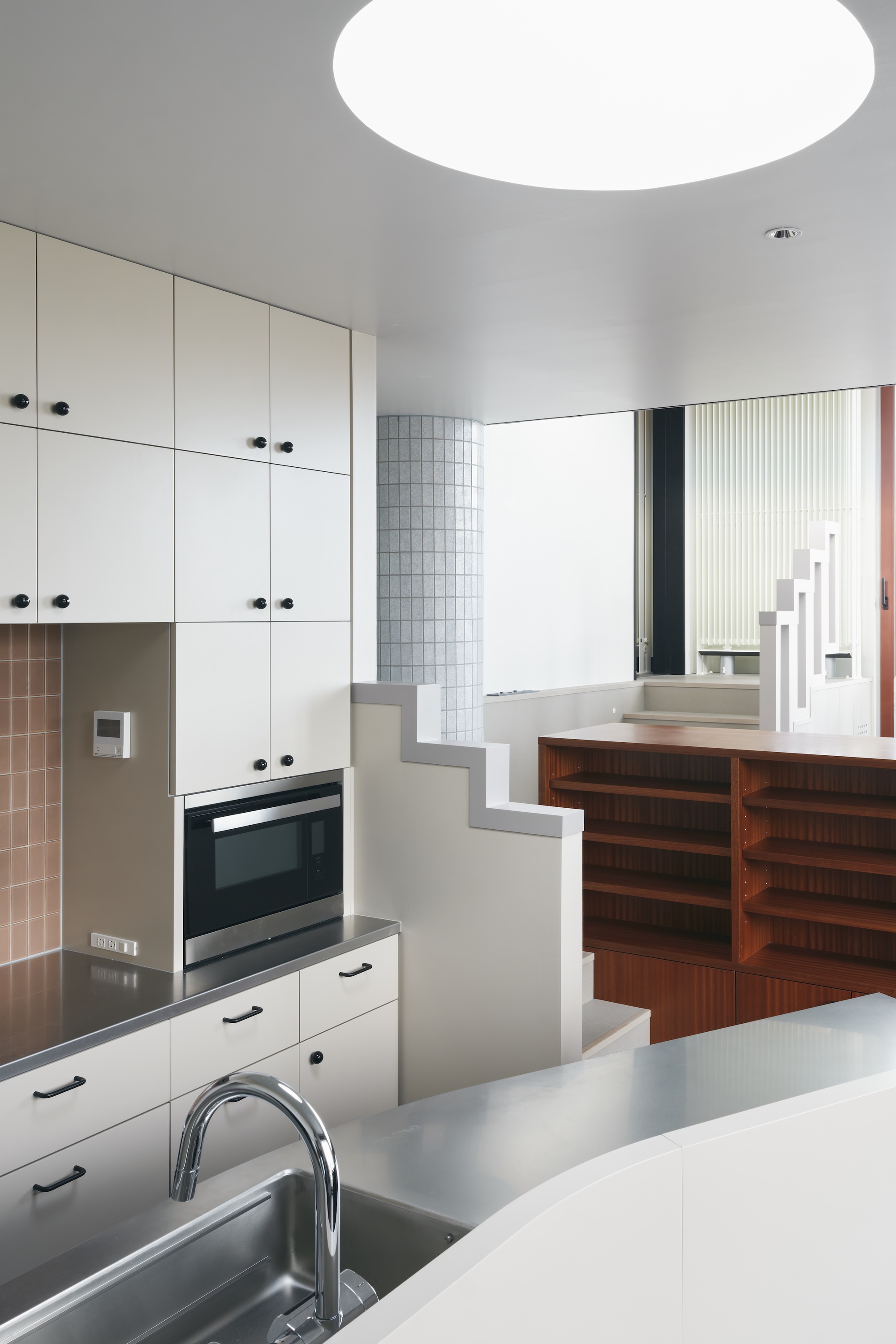 Kitchen with white cabinetry and steel counter looking toward a stepped partition and timber shelving under a round skylight