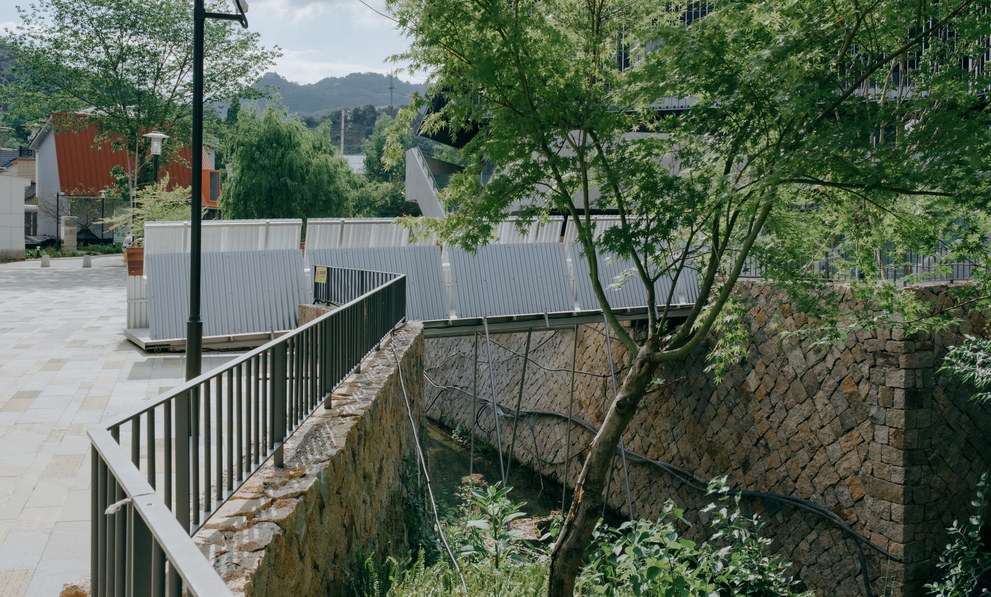 Metal railing and stacked stone retaining walls along a narrow channel, with corrugated panels beyond and a maple branch overhead