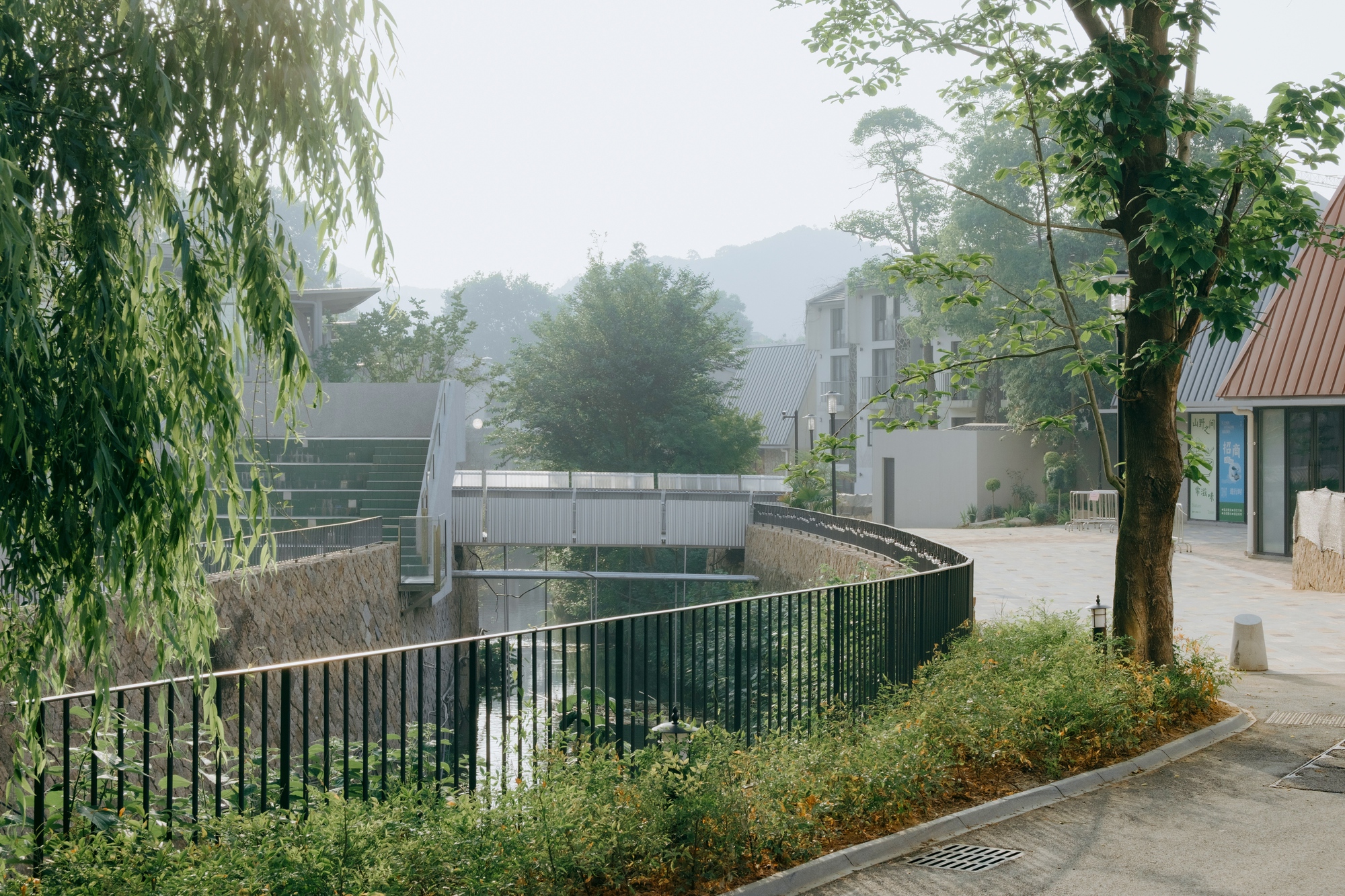 Curved dark railing above a stone-lined stream, with a corrugated footbridge in soft morning haze behind a willow and a tree trunk