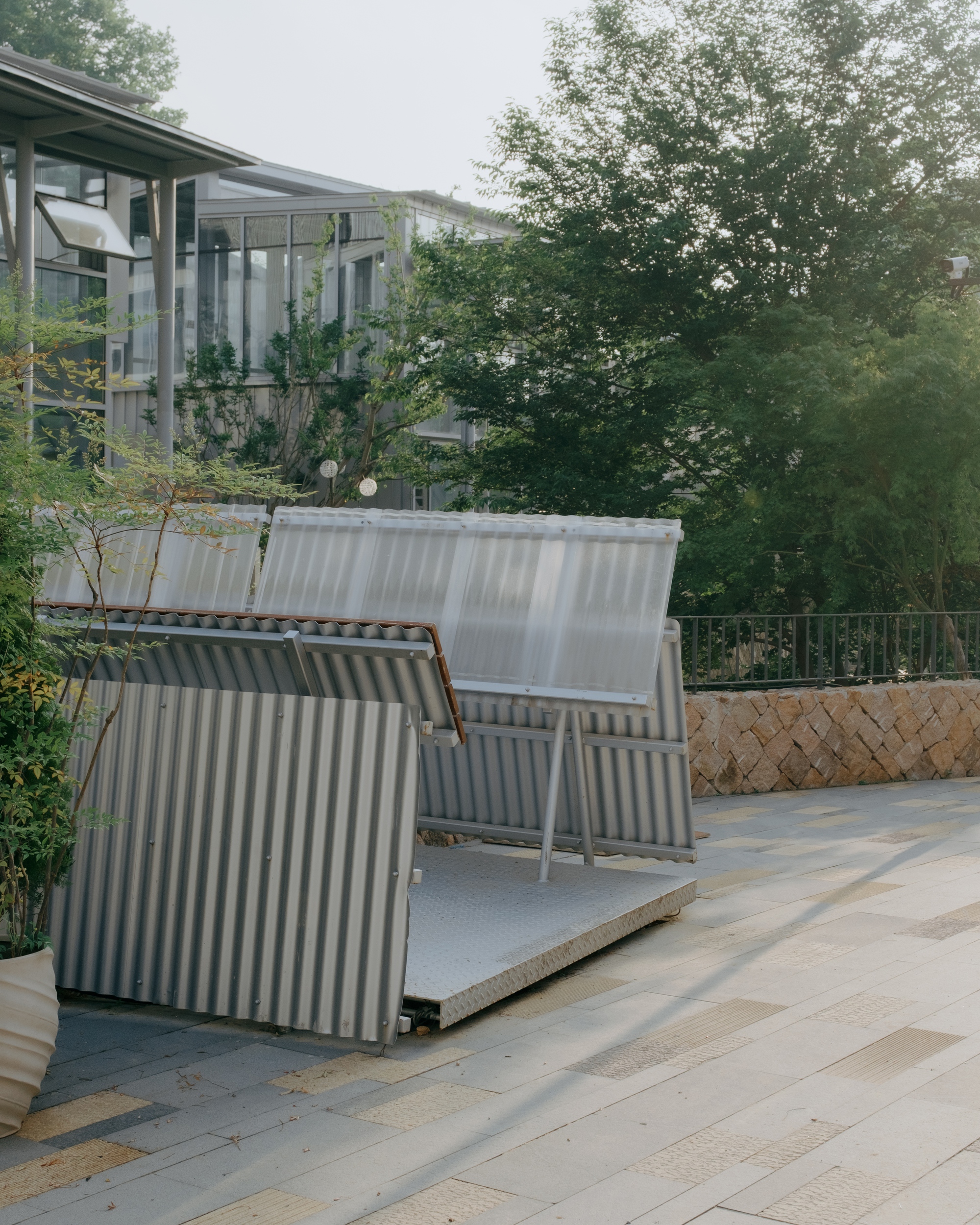 Tilted corrugated metal and translucent panels on a checker-plate platform in a paved courtyard, lit by low evening sun