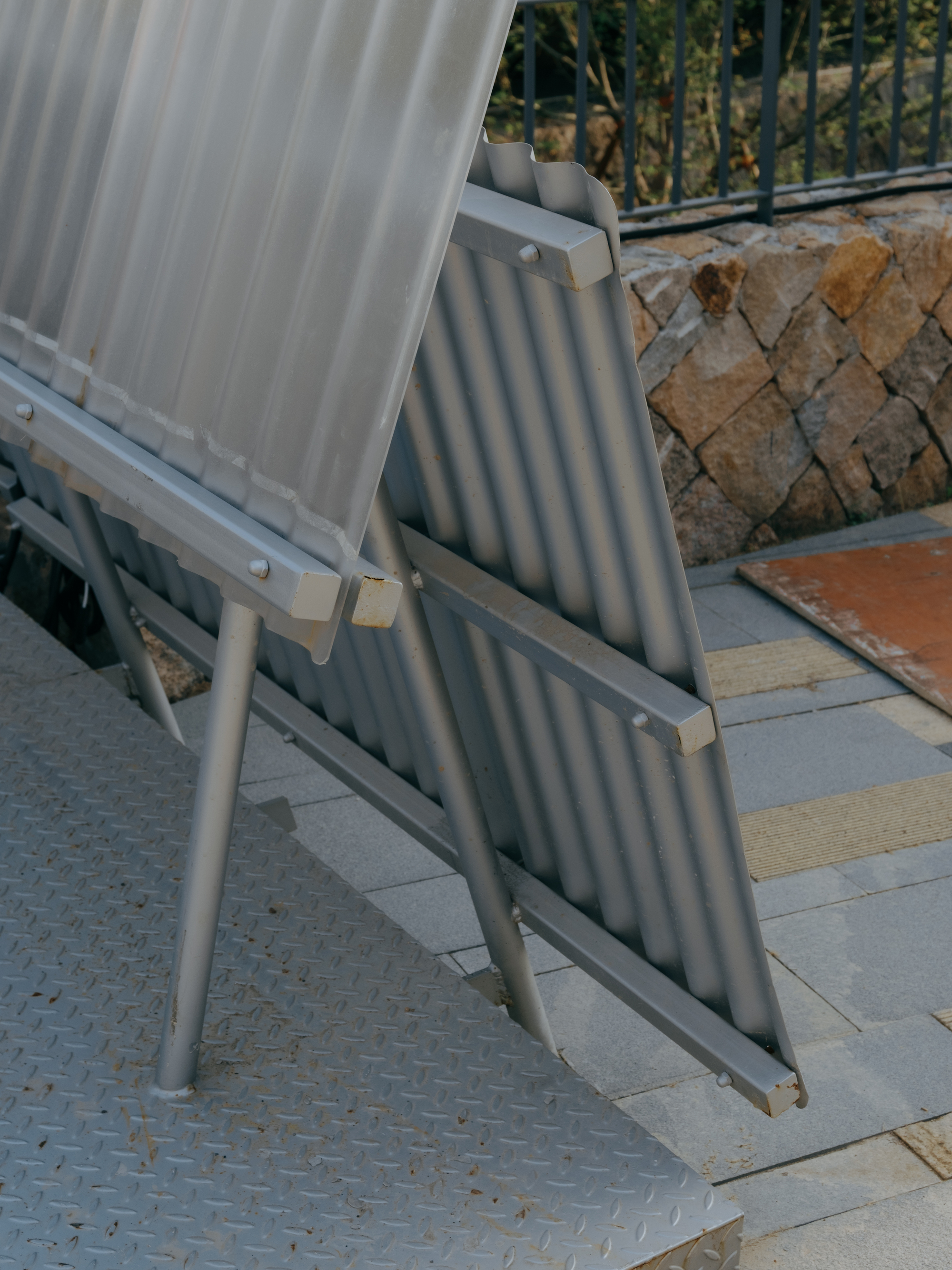 Close-up of bolted steel angle frames and tubular legs holding corrugated panels above a diamond-plate platform, with a stone wall behind