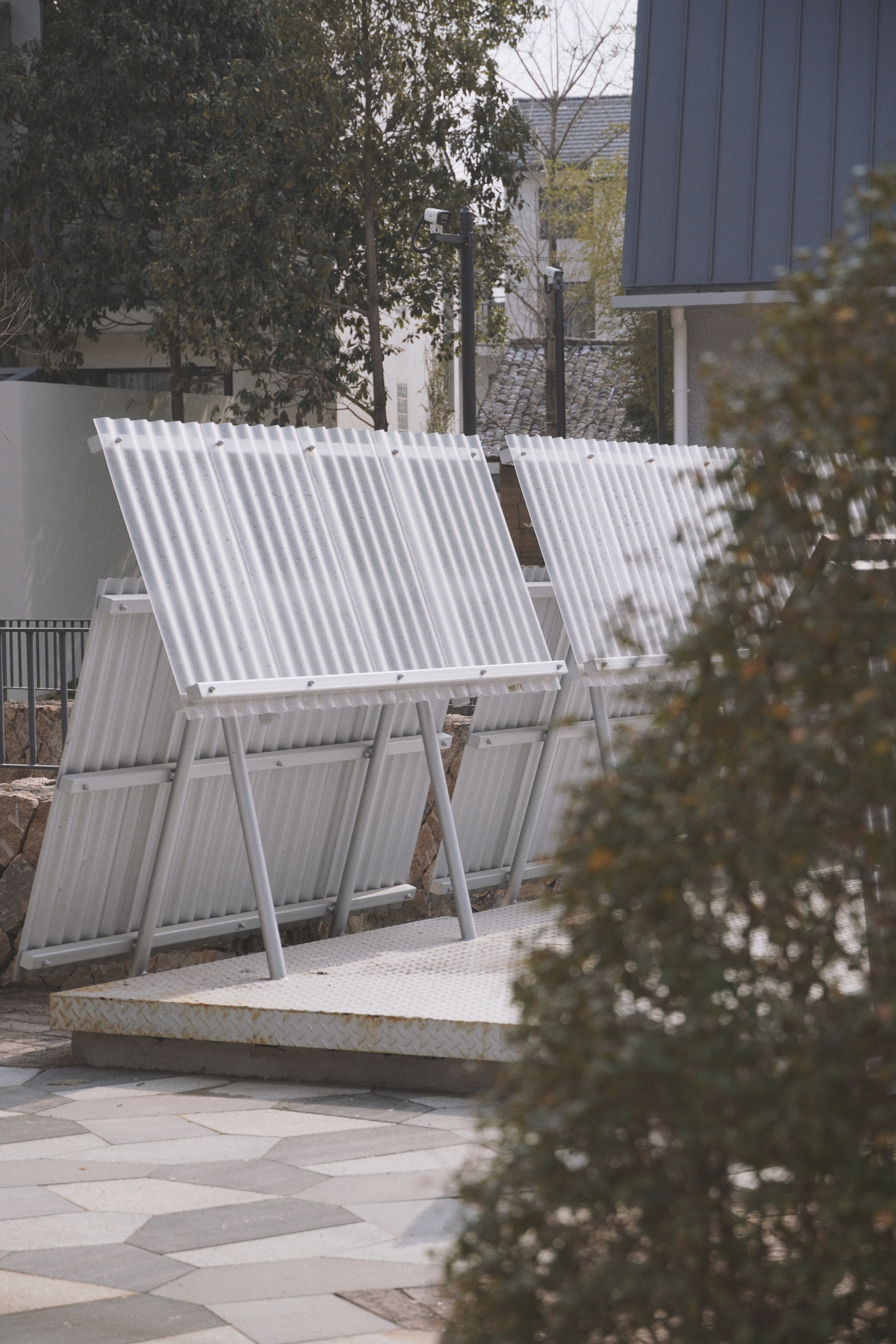 Row of angled translucent corrugated panels on slim steel legs over a checker-plate base, seen past a blurred shrub in soft daylight