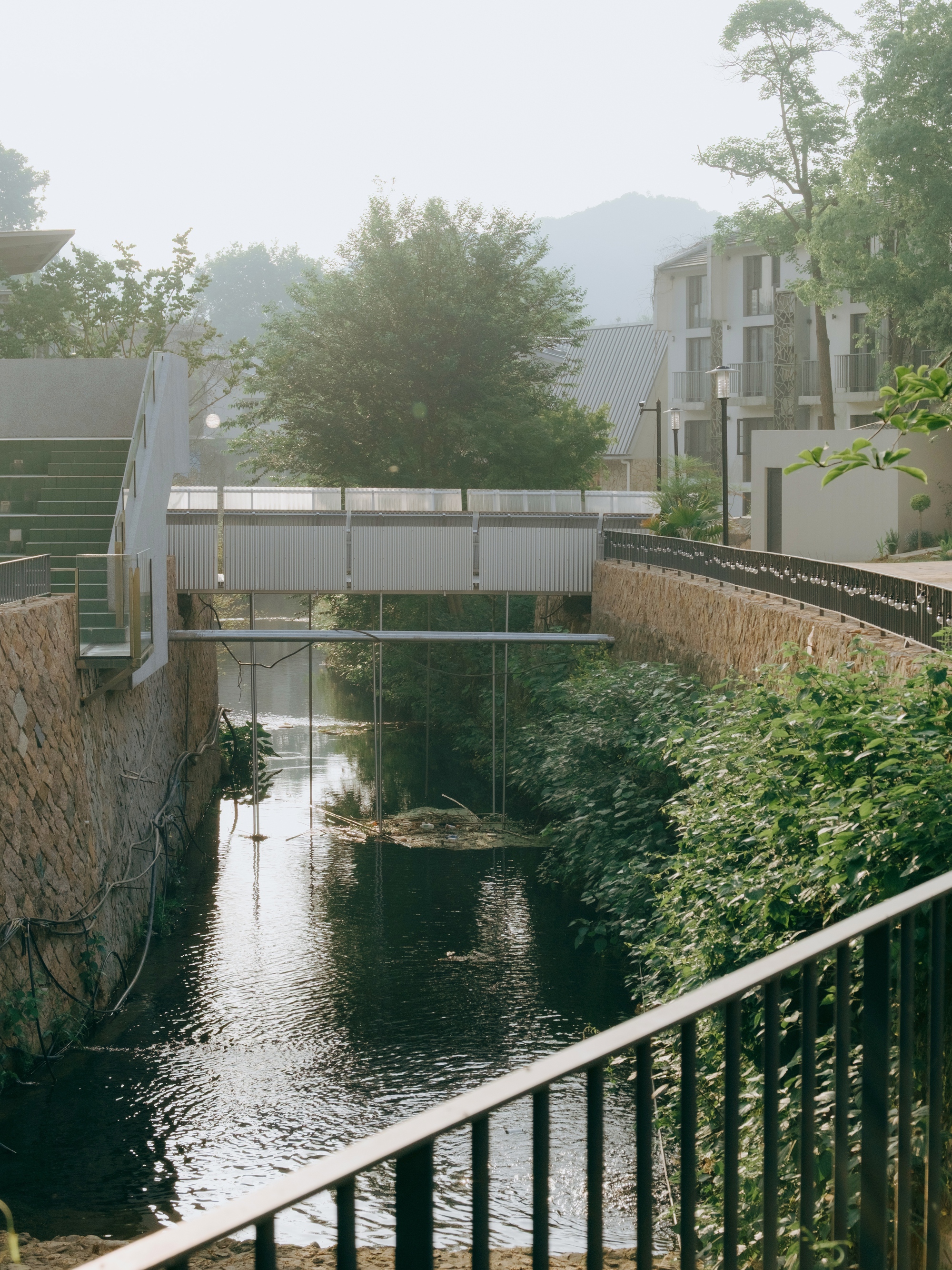 Corrugated metal footbridge spanning a narrow stream between stone retaining walls, with trees and a hillside in morning haze
