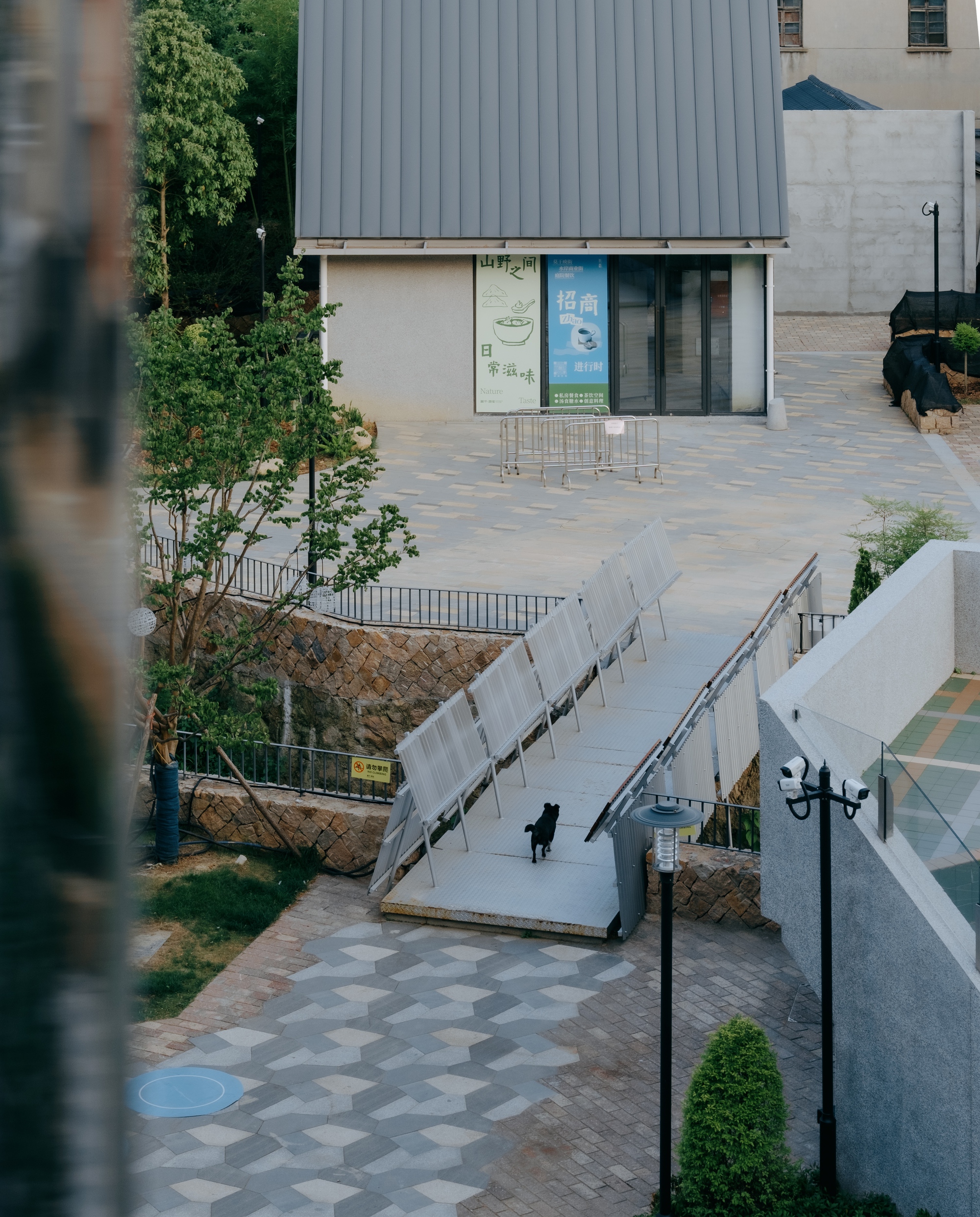 Elevated view of a metal ramp with tilted corrugated panels leading to a small gabled kiosk, a dog crossing in front