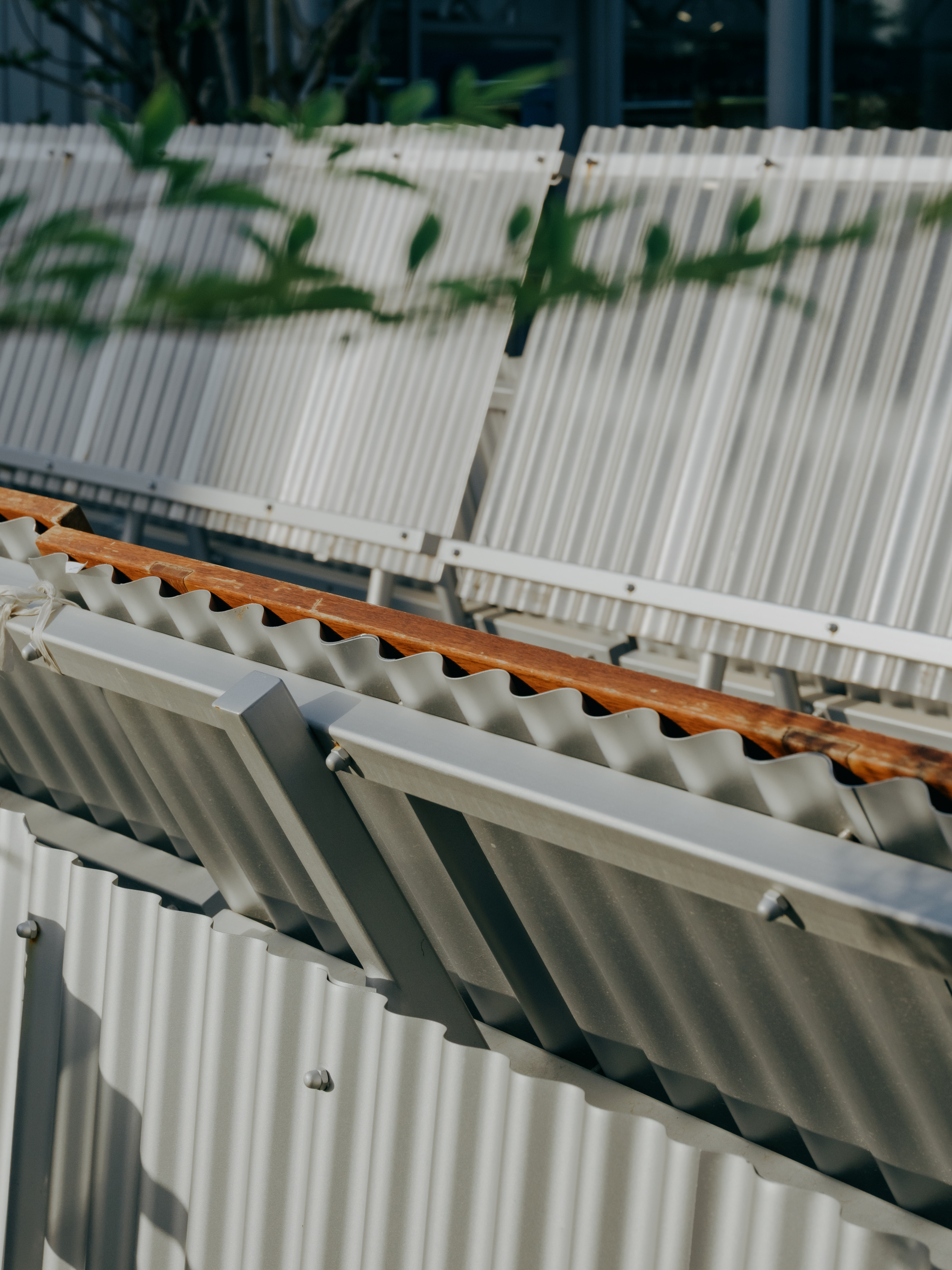 Close-up of corrugated aluminium panels tilted on steel frames with a timber rail, leaf shadows falling across the surface