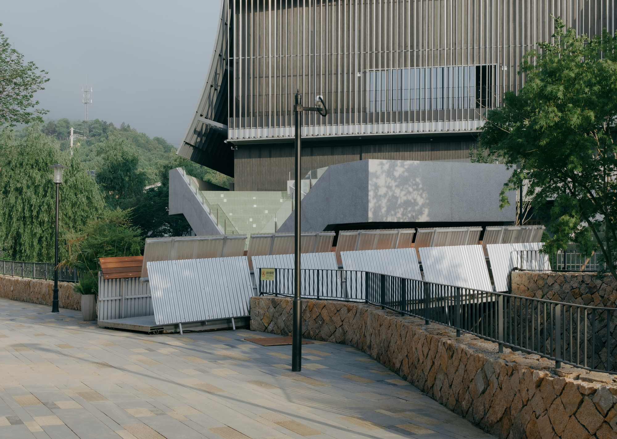 Paved plaza edged by a curved stone wall, with tilted corrugated panels below a slatted, curving building facade in hazy light