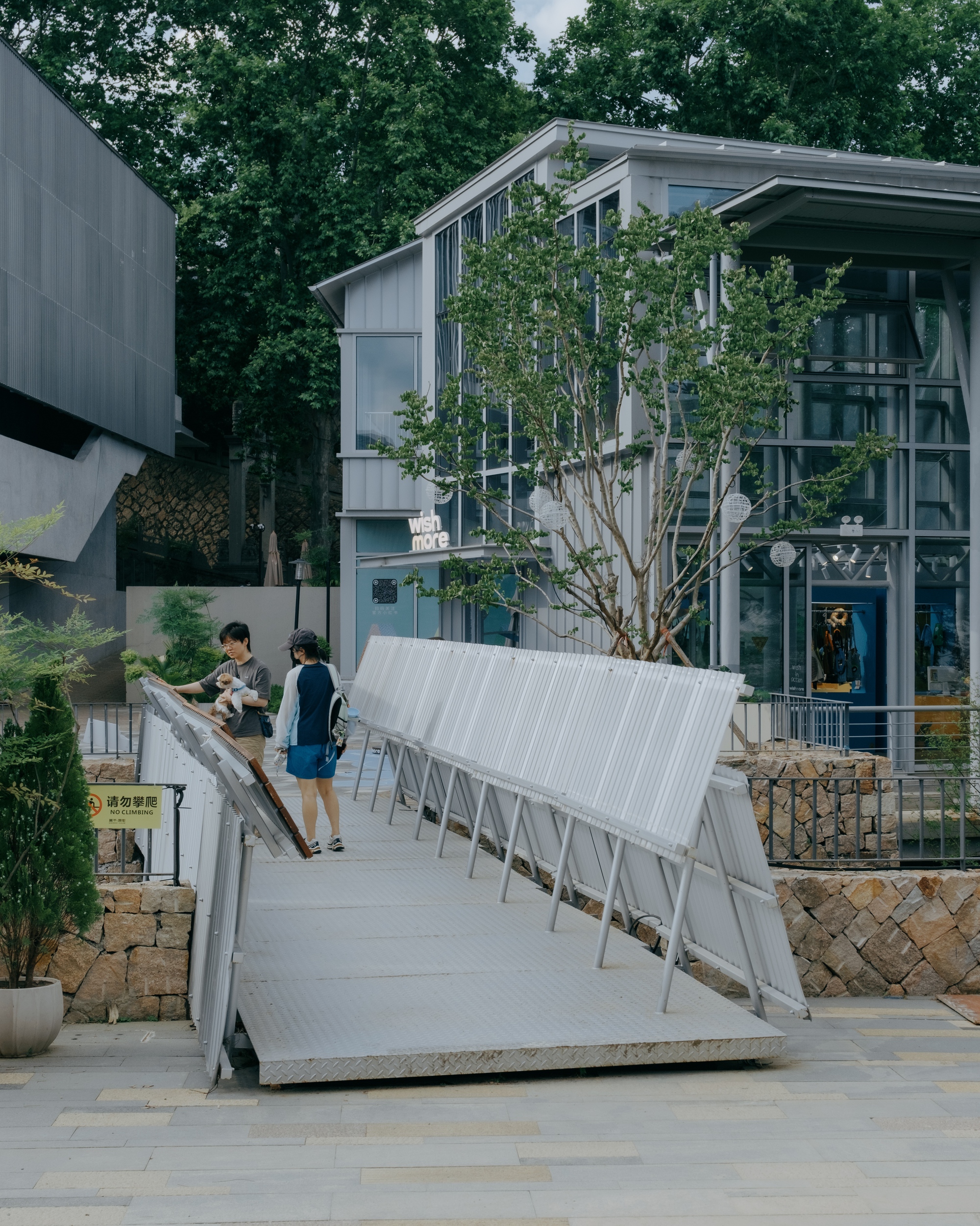 Two visitors walking up a ramp lined with tilted corrugated panels toward a glass-fronted pavilion, a young tree in front