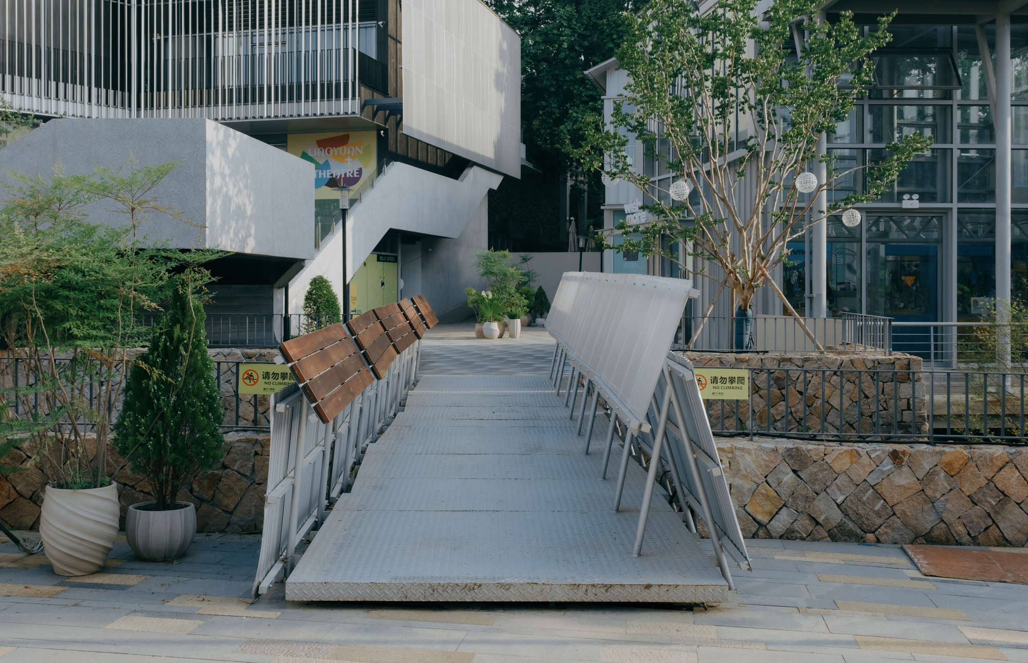 Straight metal ramp between tilted corrugated panels and timber seat rails, with potted conifers and a concrete stair behind