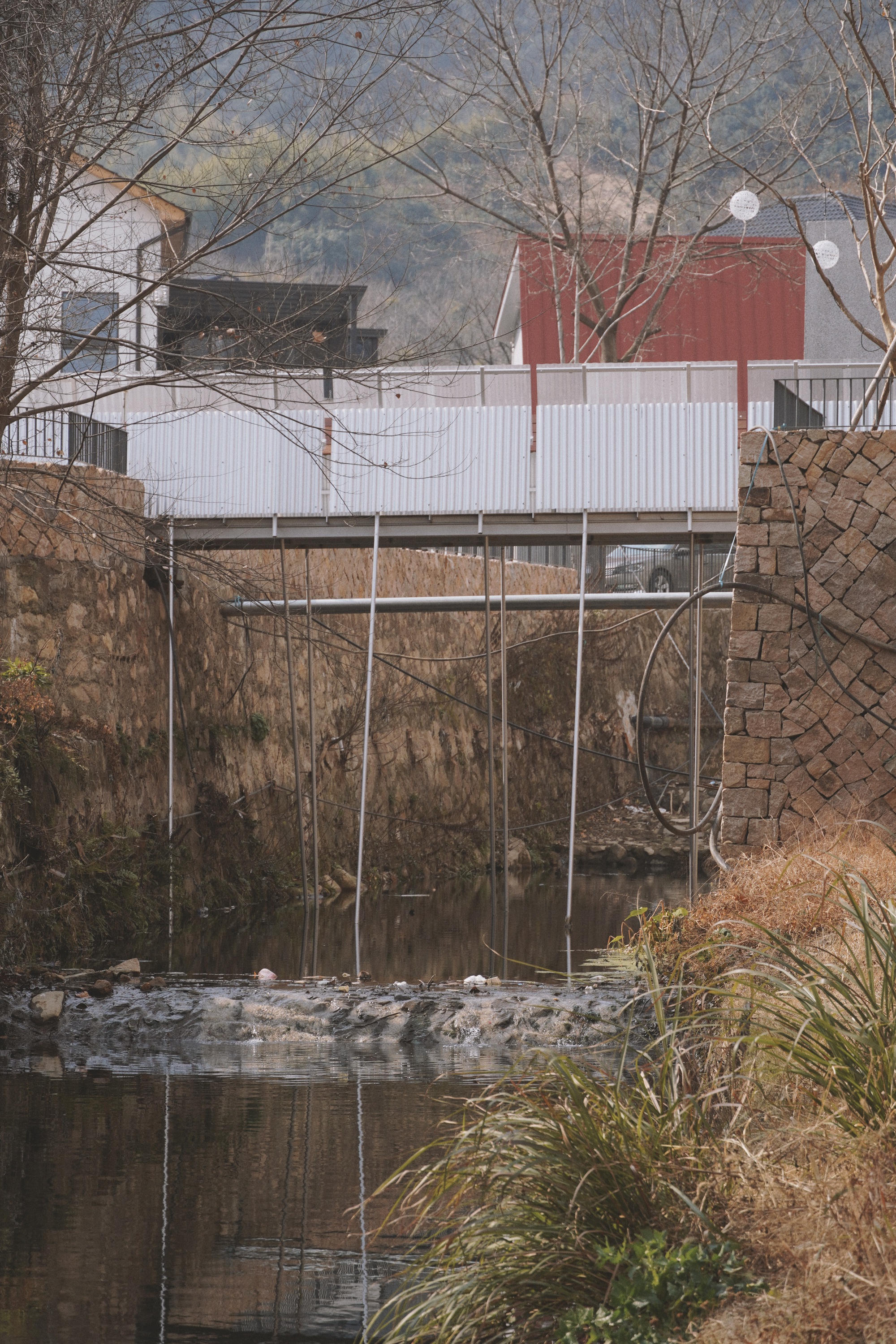 Corrugated metal footbridge on thin steel rods spanning a stone-walled stream, seen through bare winter branches