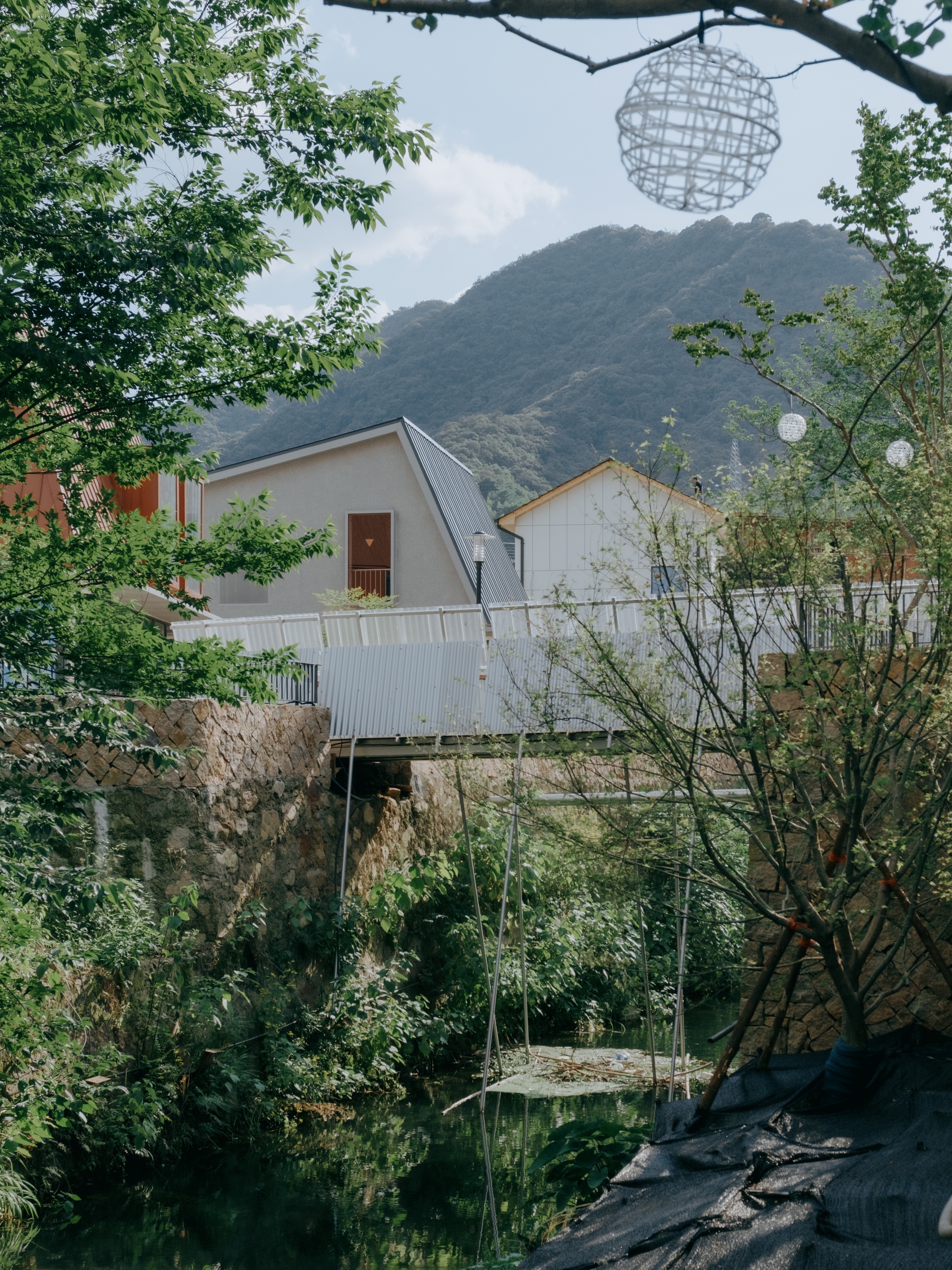 Footbridge with corrugated metal balustrade crossing a stream below gabled houses and a green mountain in summer light