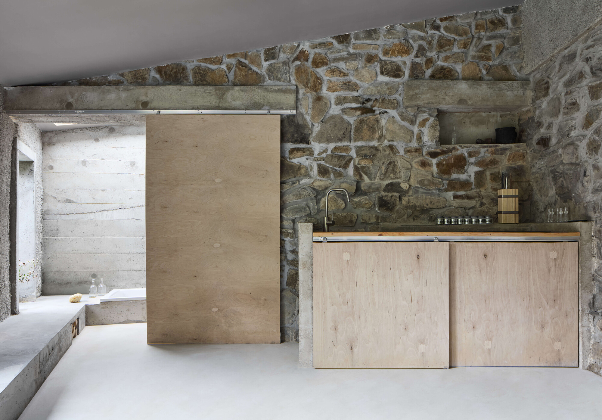 Rubble stone wall with plywood sliding door and cabinet doors above a steel sink counter, lit by soft daylight
