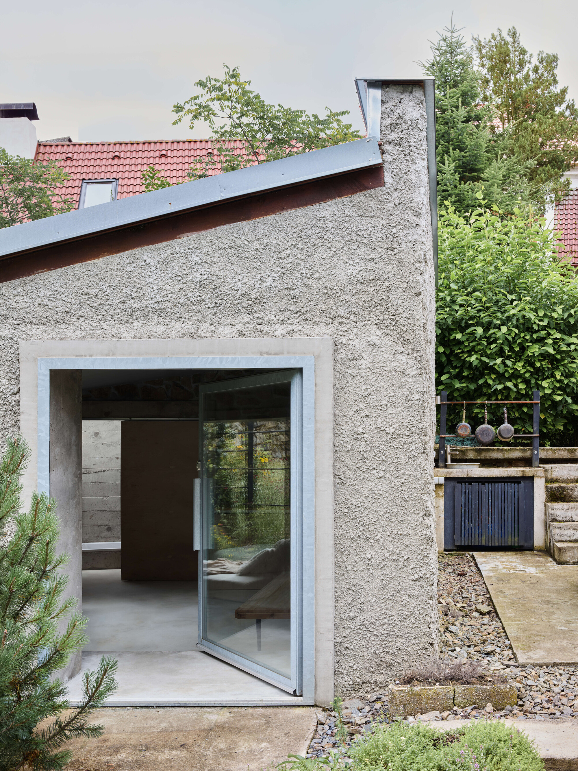 Rough plastered gable end with a steel-framed pivot glass door opening onto a gravel path and a pine sapling