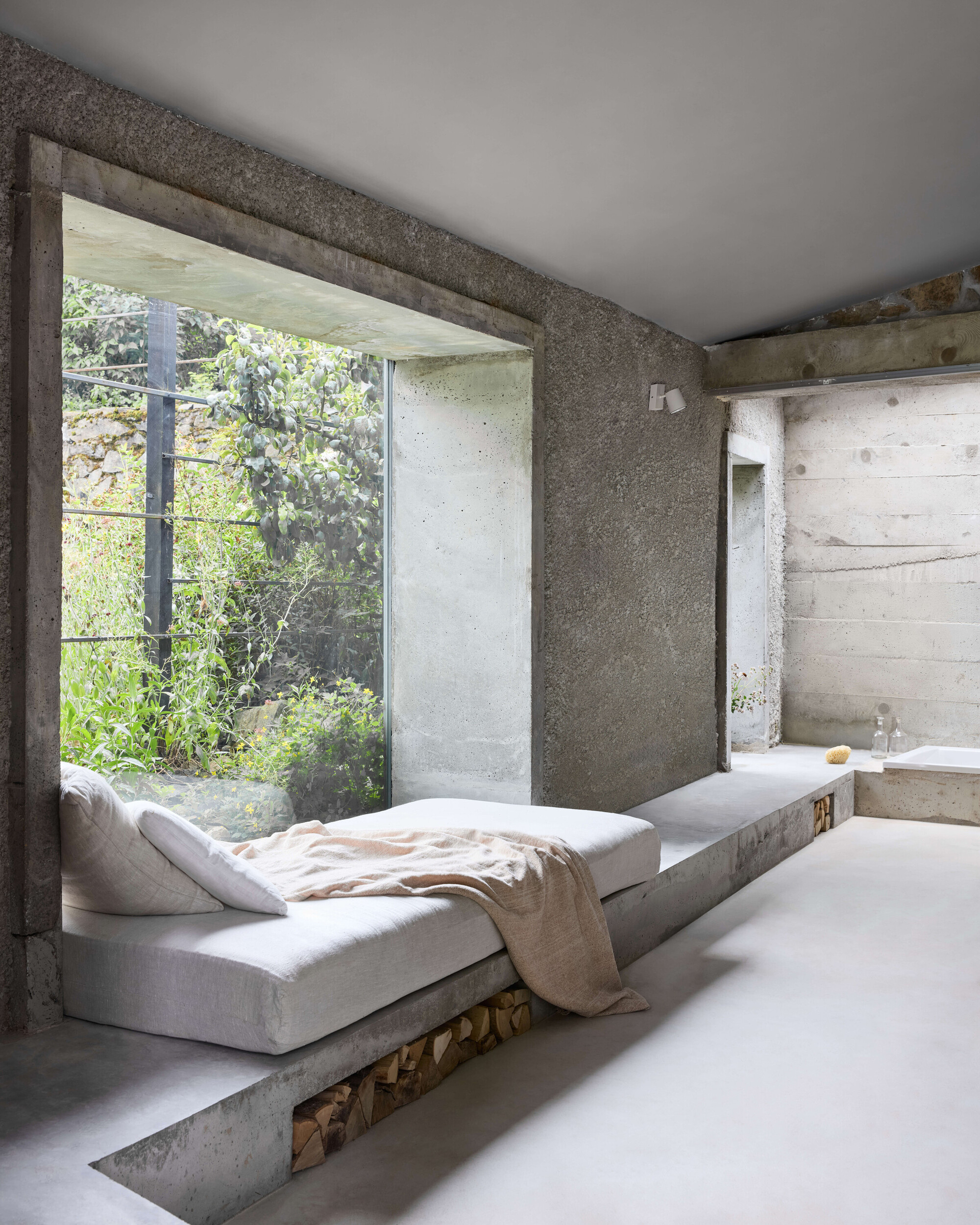 Concrete daybed platform with firewood storage beneath, facing a large framed window onto a green garden