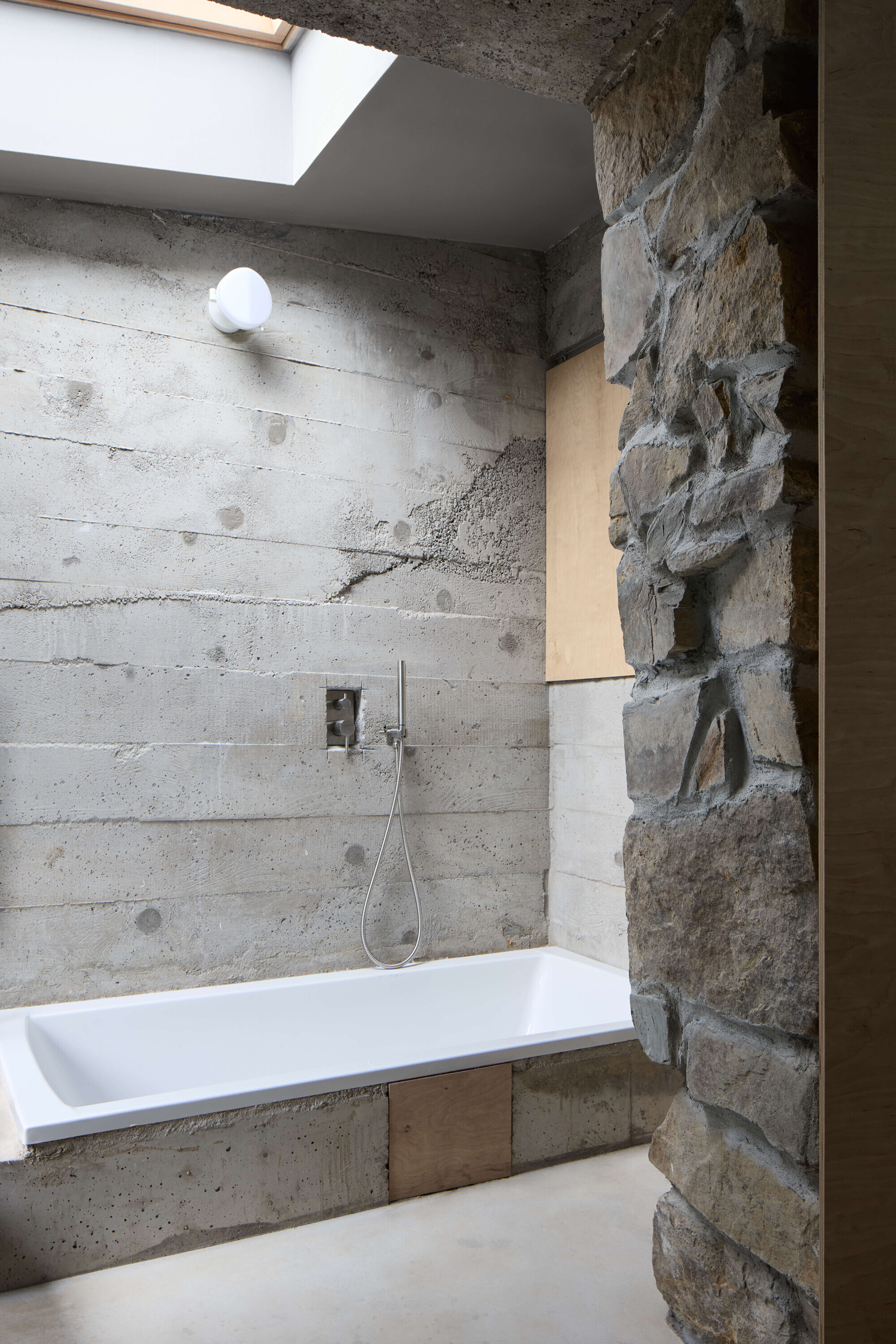 Built-in white bathtub against board-formed concrete wall, with a skylight overhead and a rubble stone pier at right