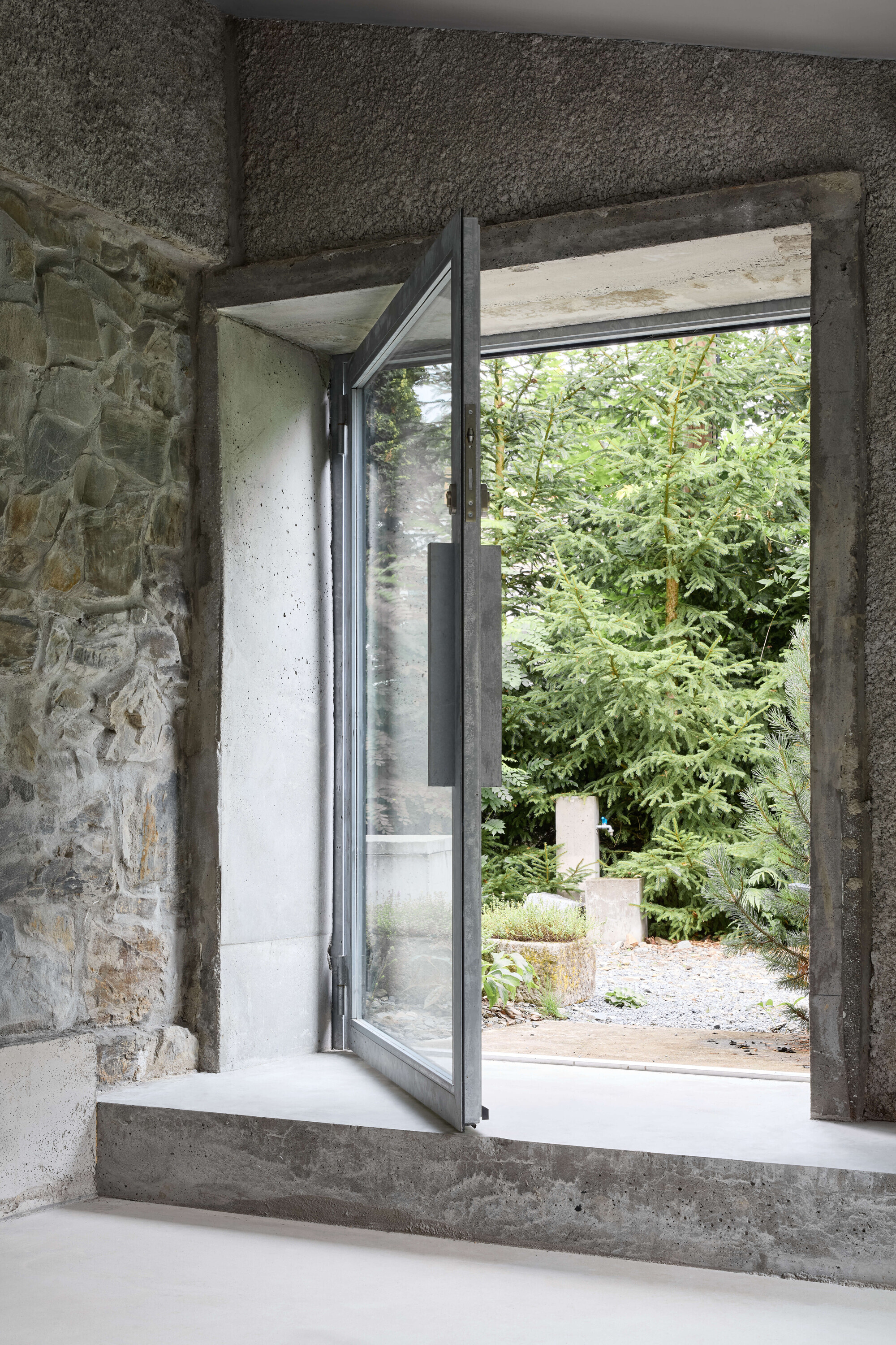Open pivot glass door set in a deep concrete reveal beside stone wall, looking out to spruce trees and gravel