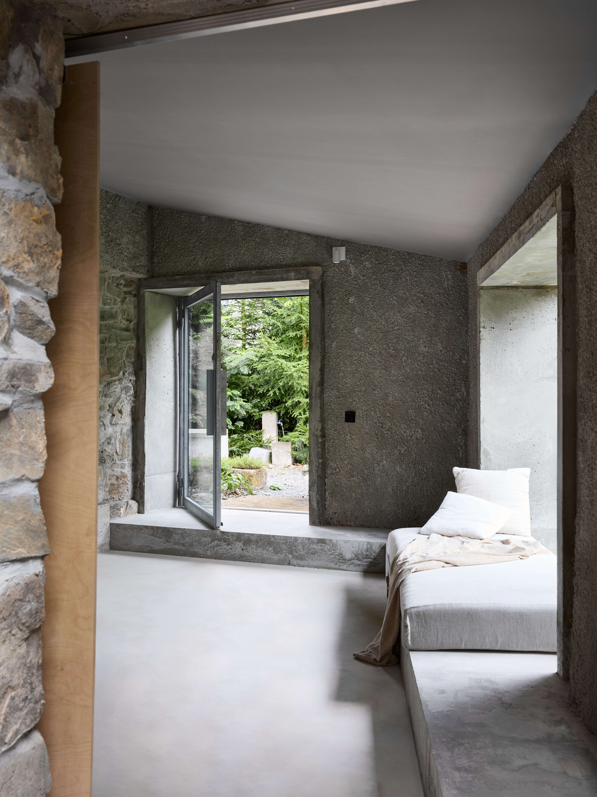 Interior with raw concrete walls, a stone jamb and a pivoting glass door opening to a green garden
