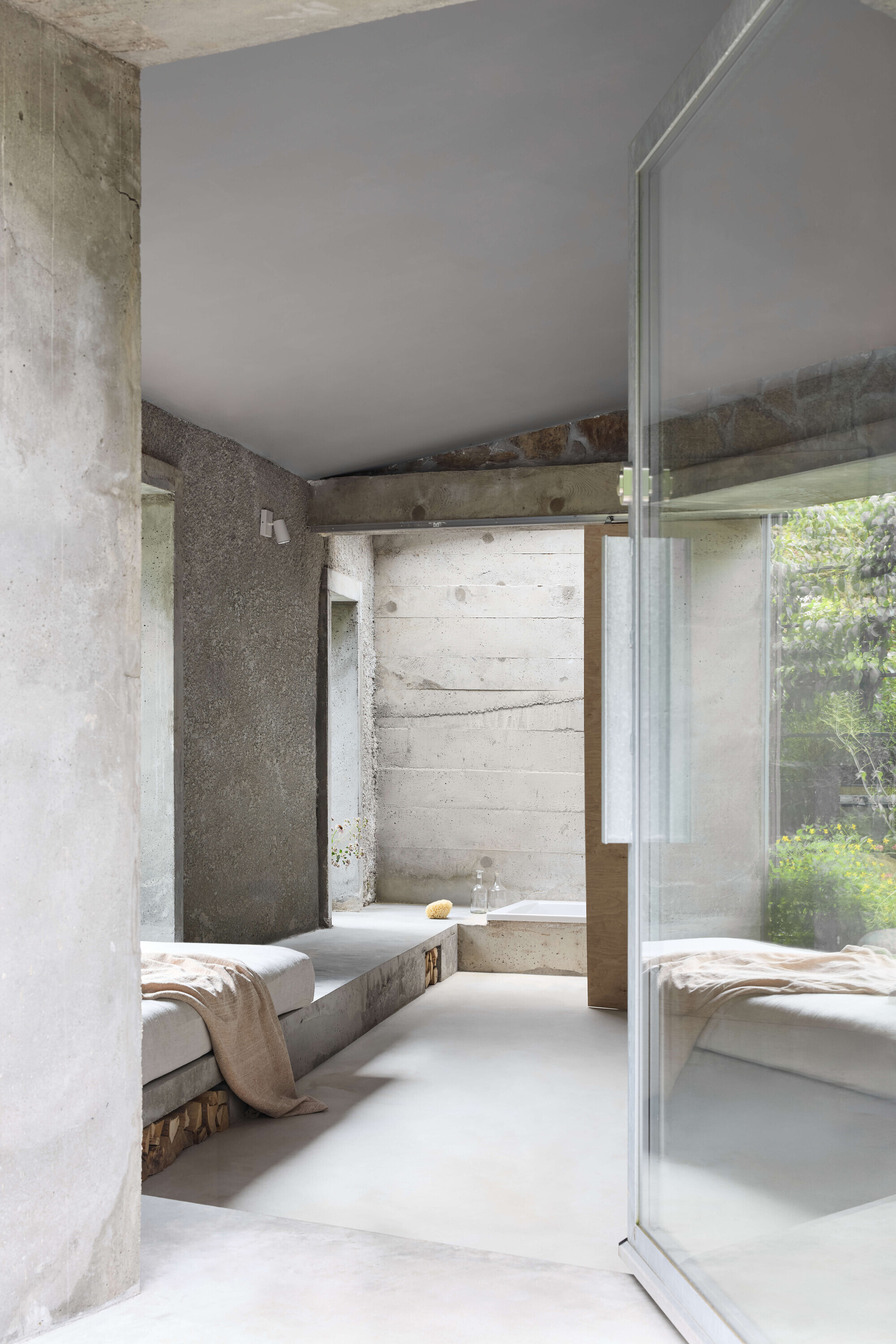 Concrete room with a raised daybed platform and floor-to-ceiling glass panel reflecting garden greenery in soft daylight