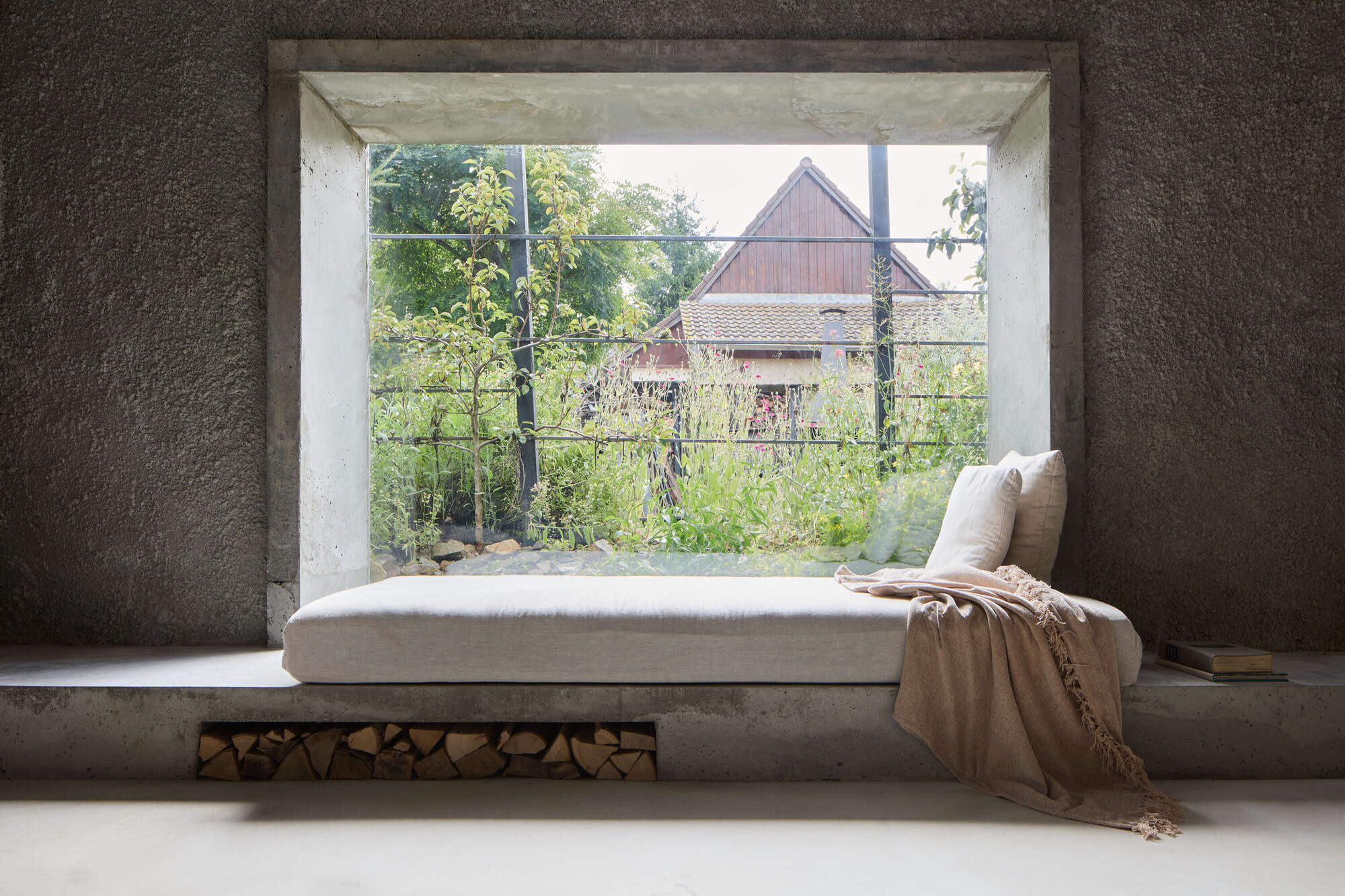 Built-in concrete daybed with linen cushions and a stacked firewood niche beneath a large window framing a gabled house