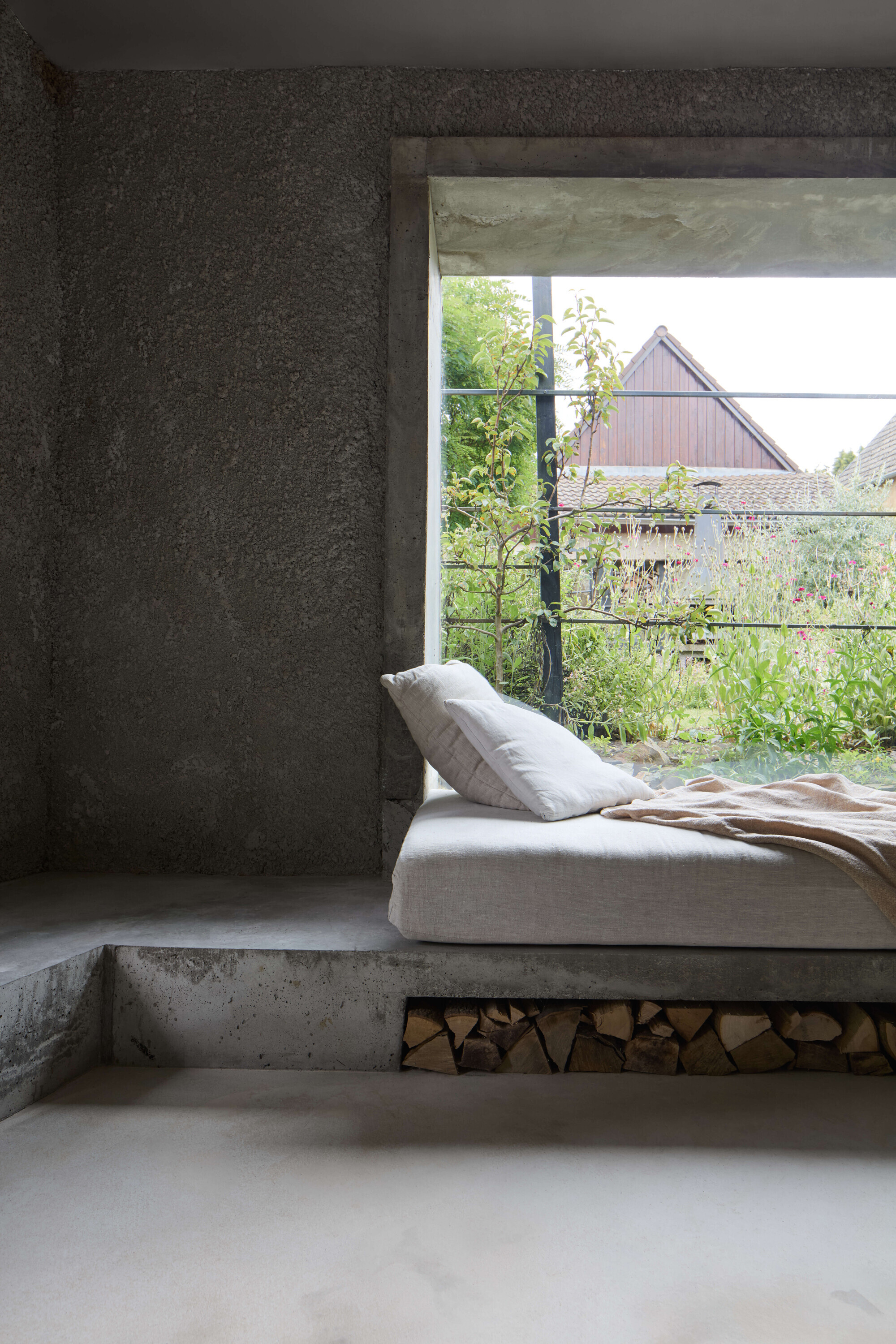 Side view of the concrete daybed platform with stacked logs below and pillows facing a window onto a wildflower garden