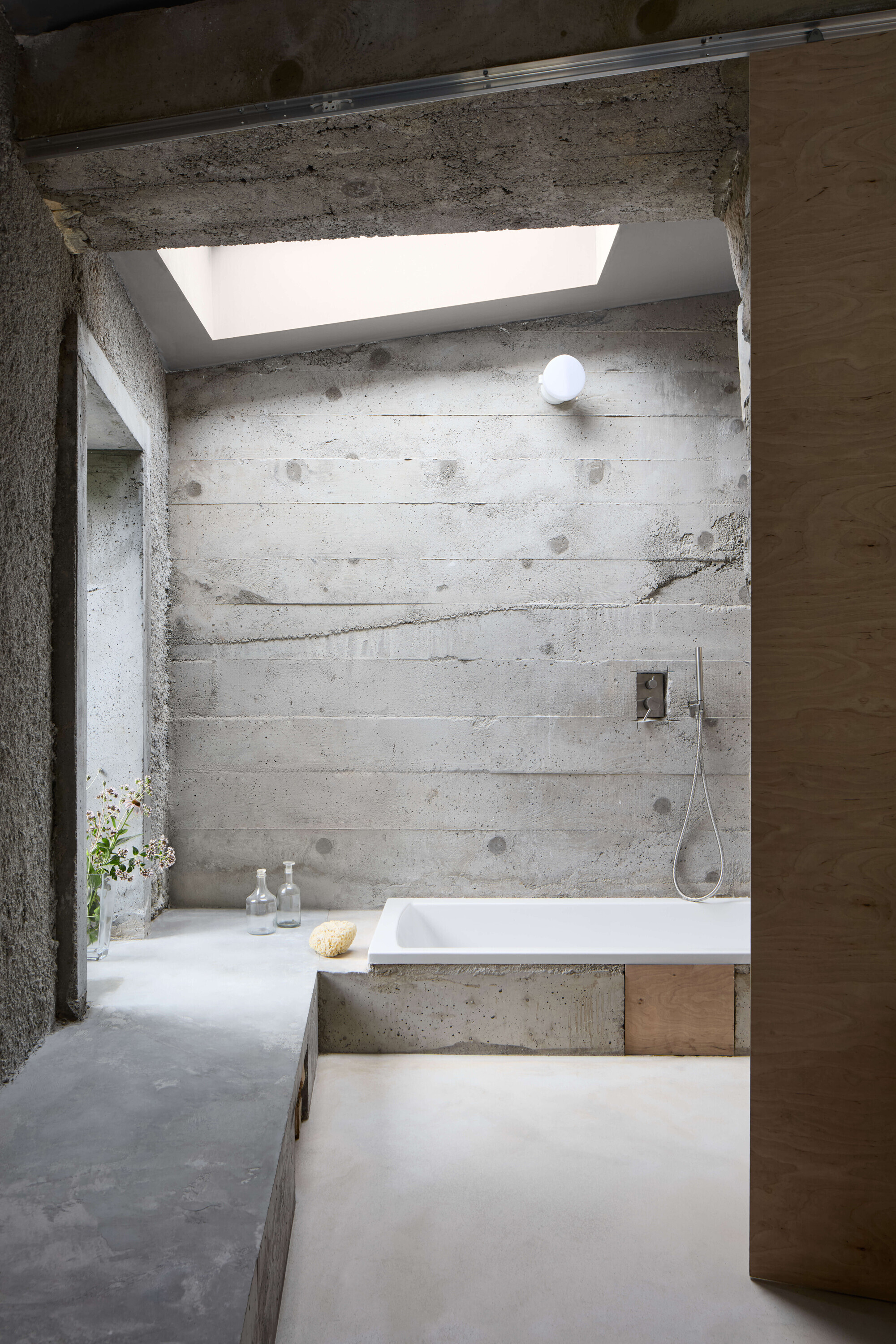Bathroom with board-marked concrete wall, a sunken white tub and skylight, with glass bottles and wildflowers on the ledge