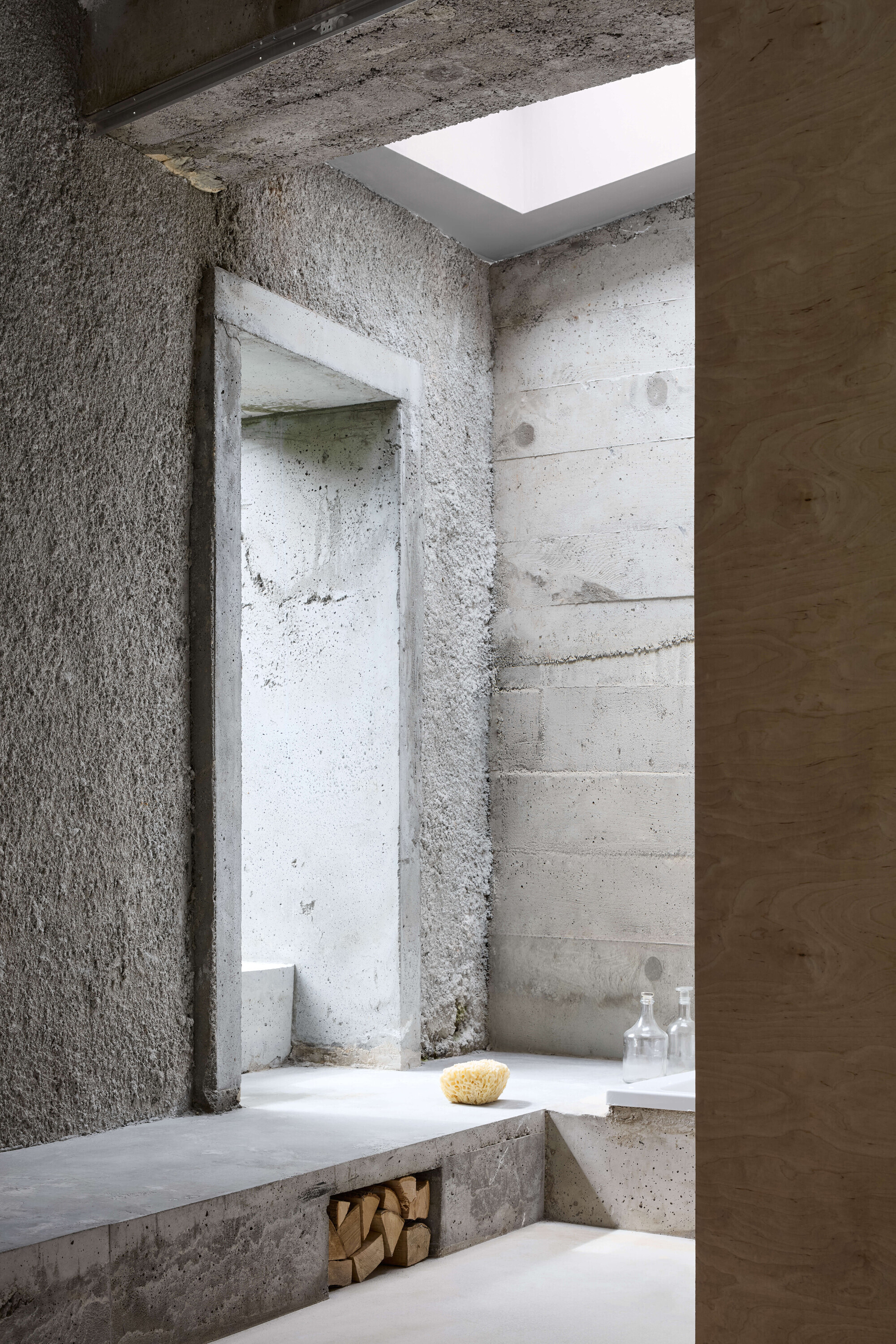 Rough concrete bathing room with a recessed doorway, stacked firewood under a ledge and a skylight casting soft daylight