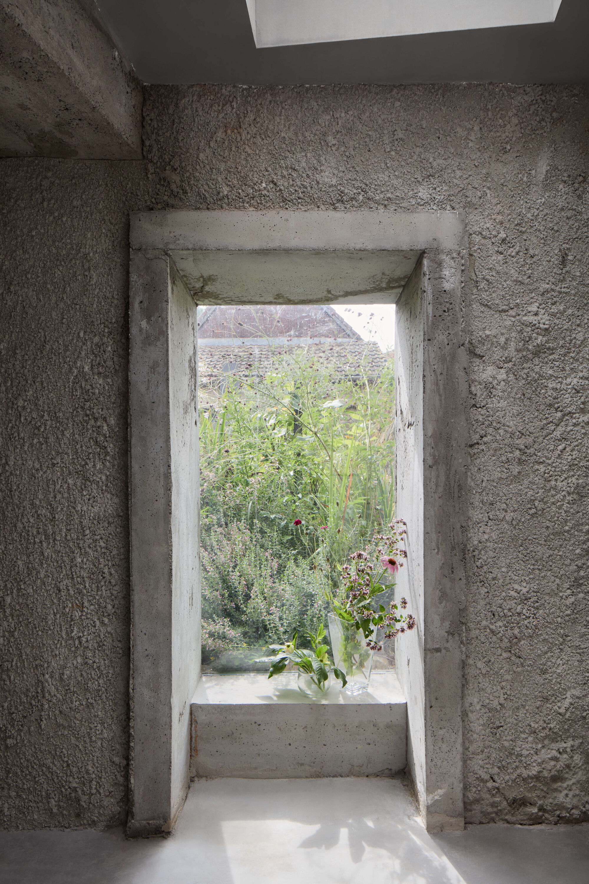 Deep concrete window reveal framing a wild garden, with cut flowers in a glass vase on the sill