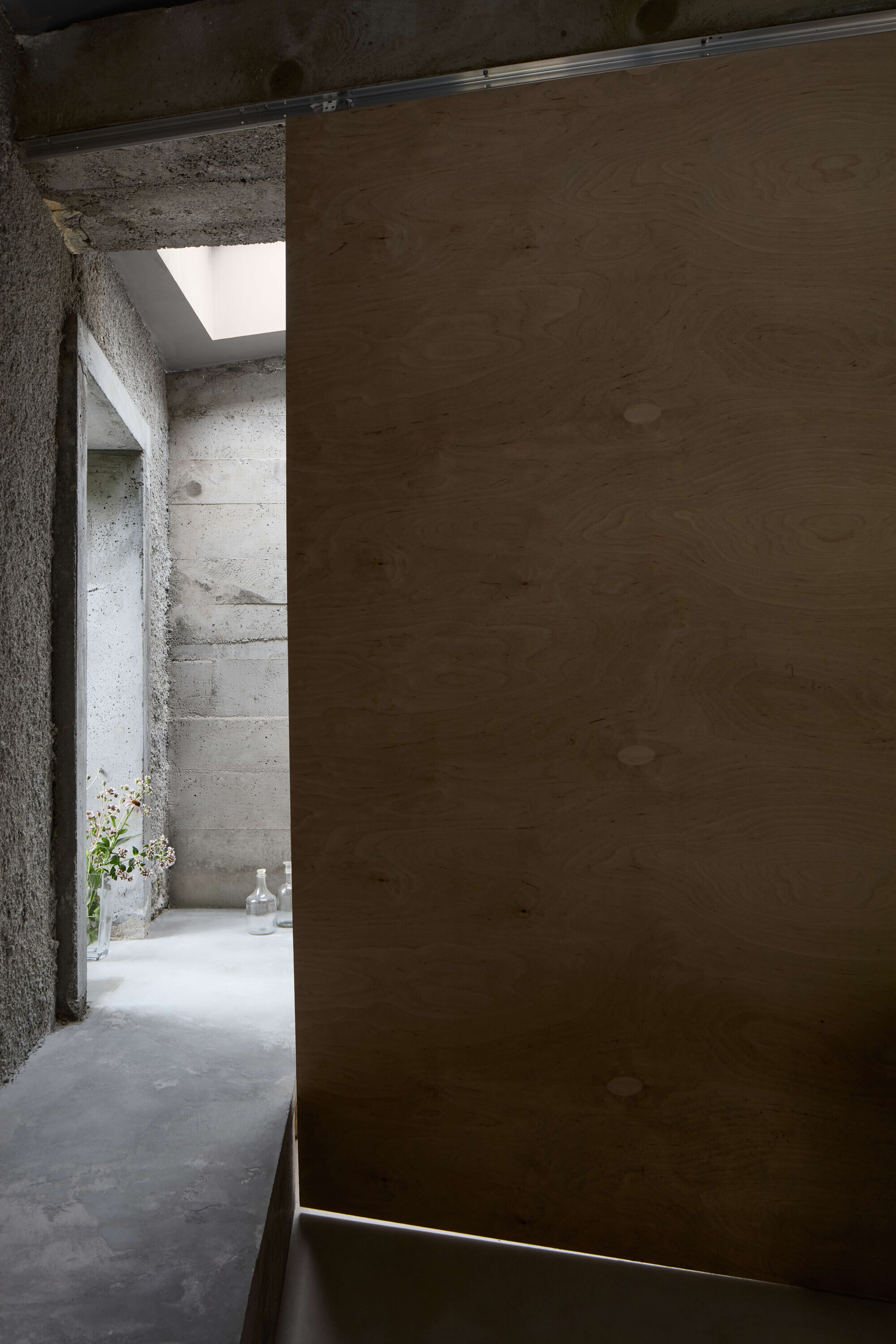 Sliding plywood panel in the foreground, with a concrete passage, glass bottles and a skylight glowing beyond
