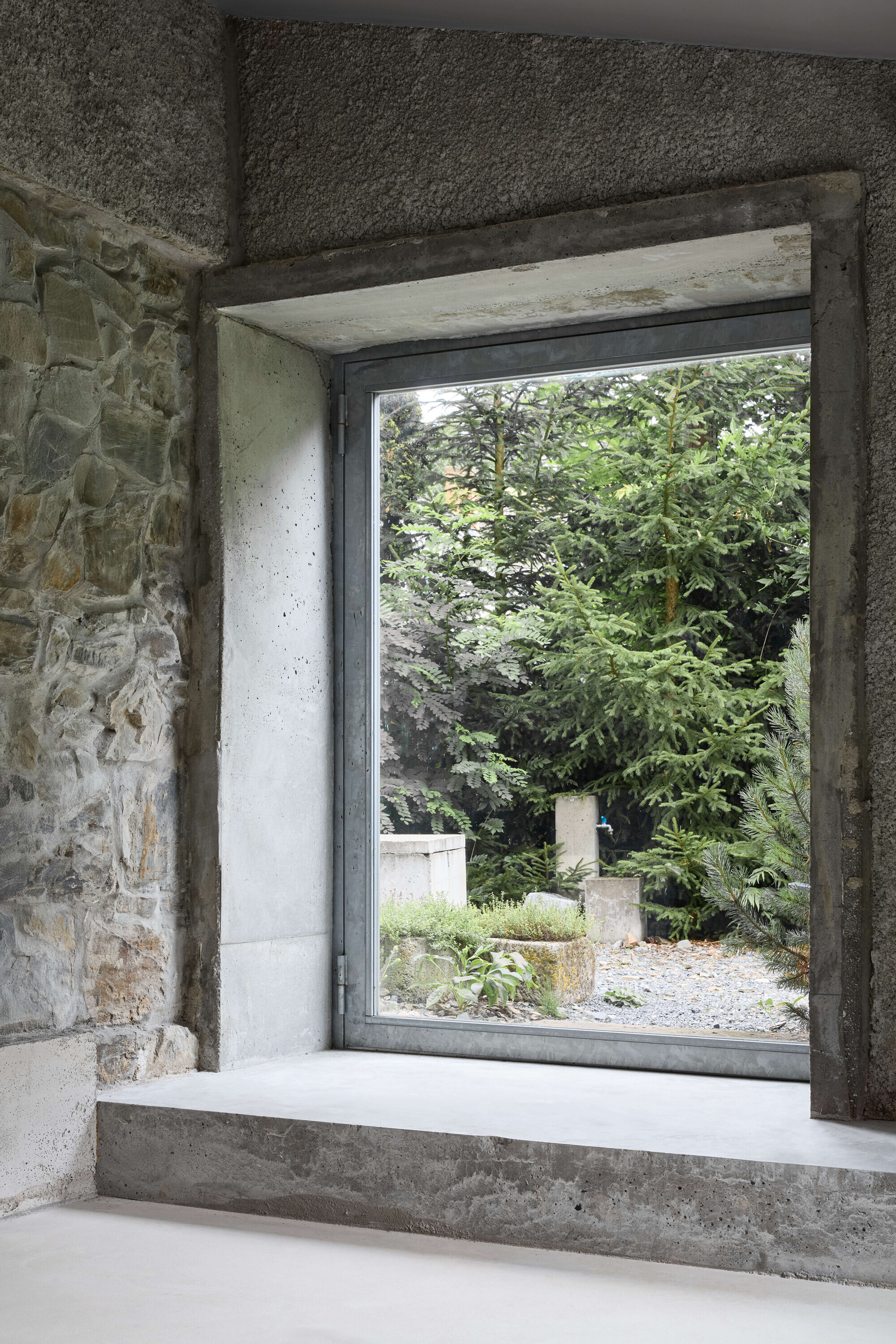 Large steel-framed window set into a concrete reveal beside exposed rubble stone, looking onto conifers and a gravel garden