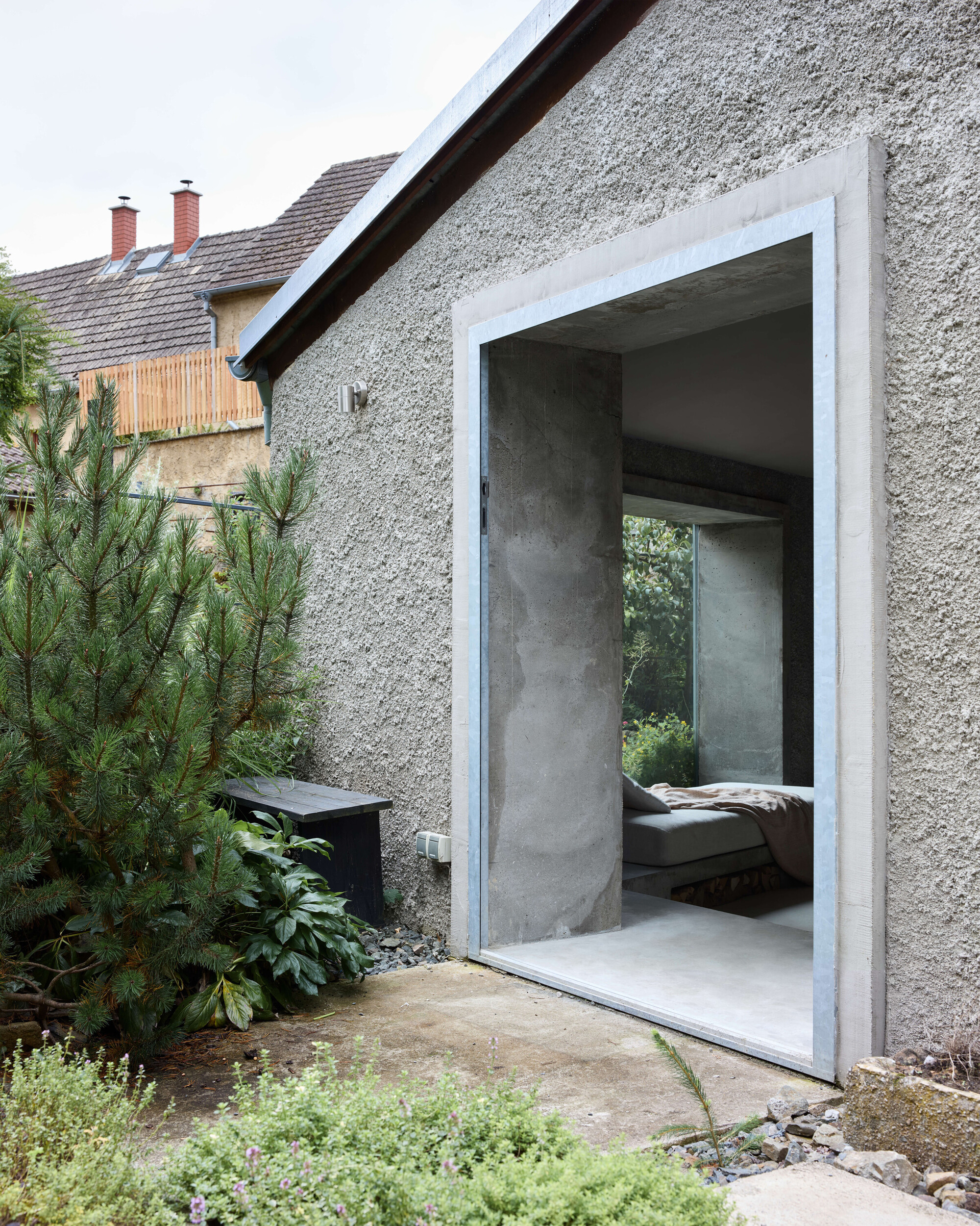 Tall concrete-framed opening in a rough plastered gable wall, showing a daybed inside and pine and thyme in the foreground