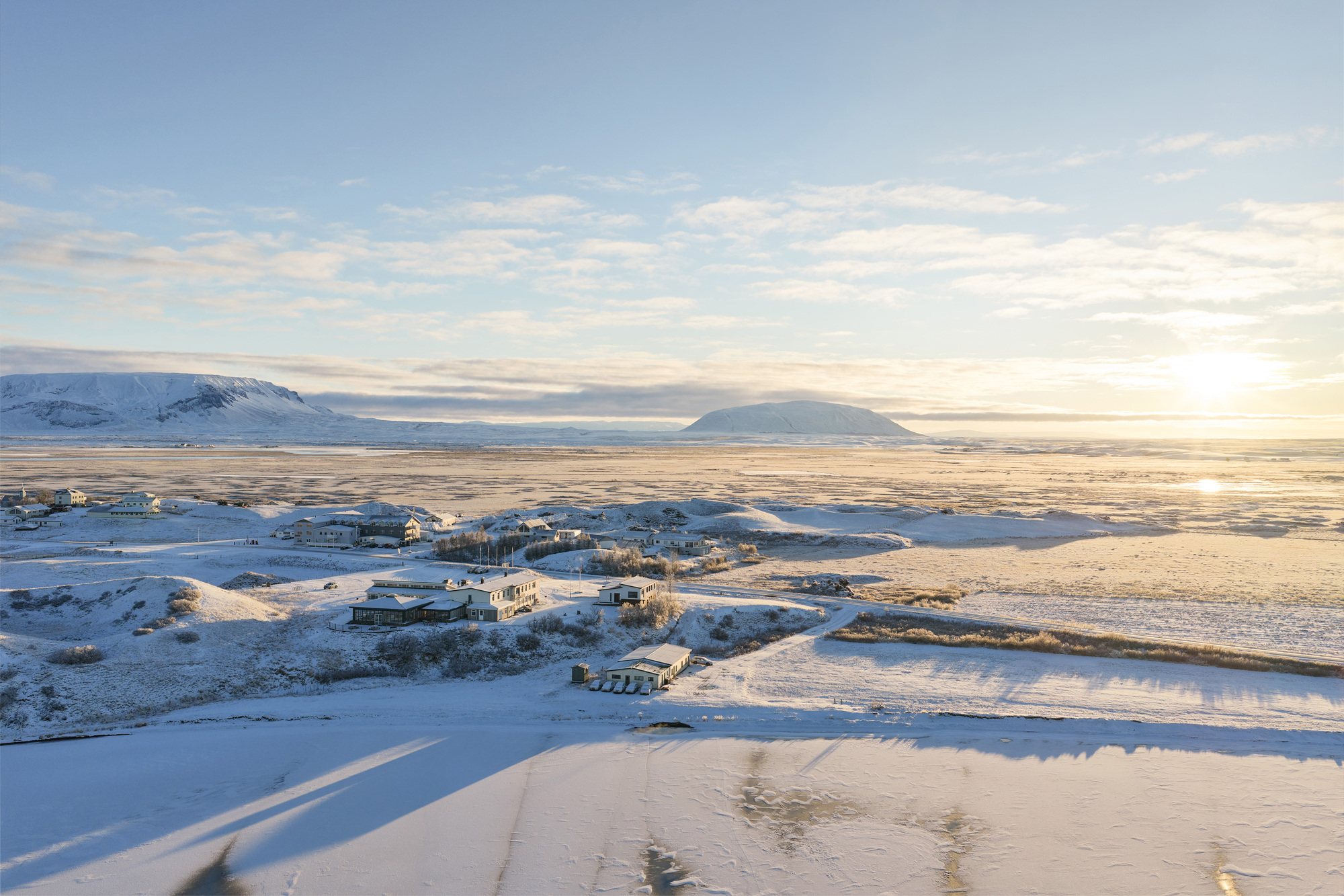 Aerial view of a cluster of low buildings on a frozen plain at sunrise, with snow-covered hills behind