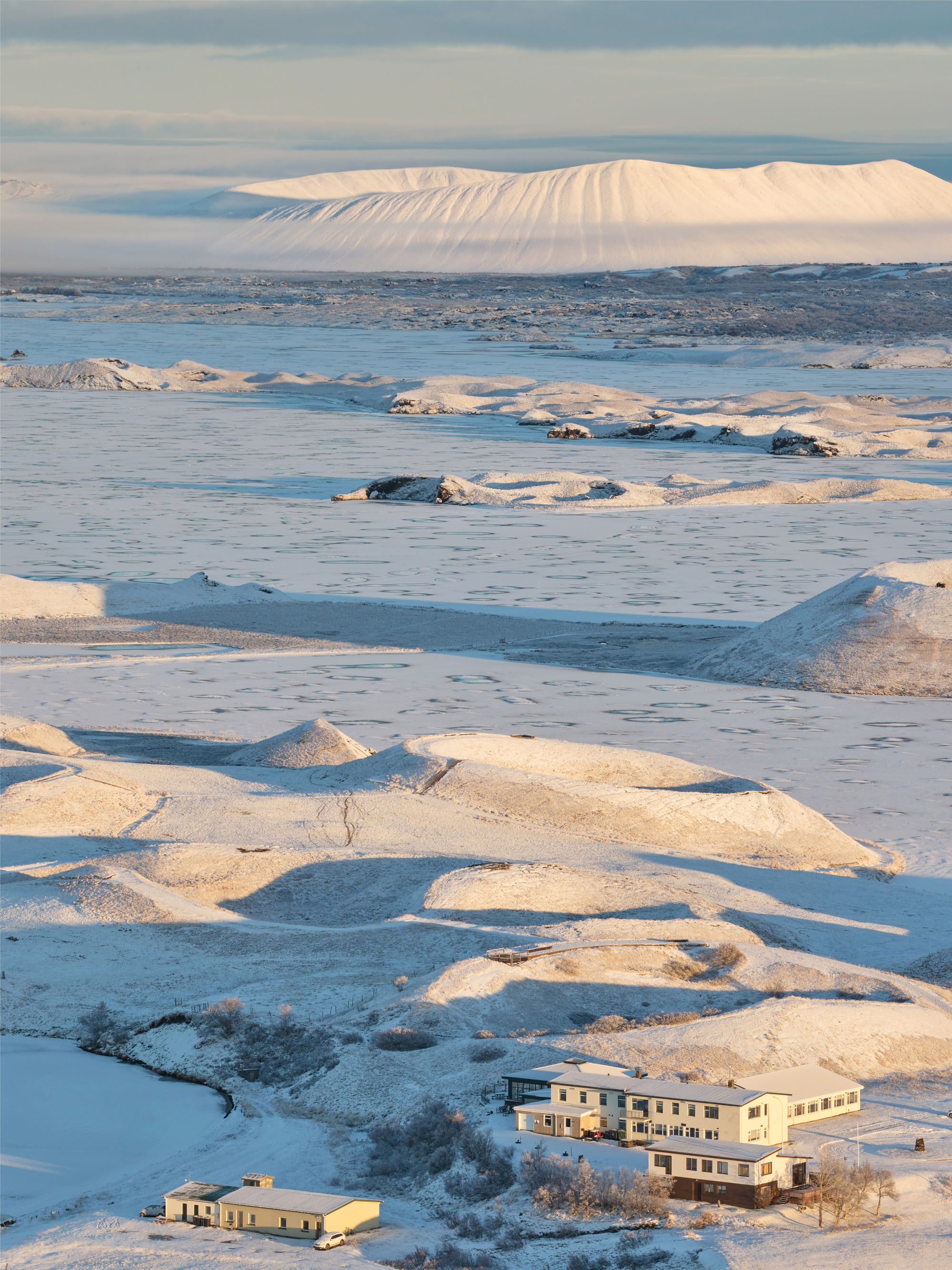 Telephoto view of a pale multi-wing building and a small outbuilding among snowy mounds, with a white crater rim beyond in low sun